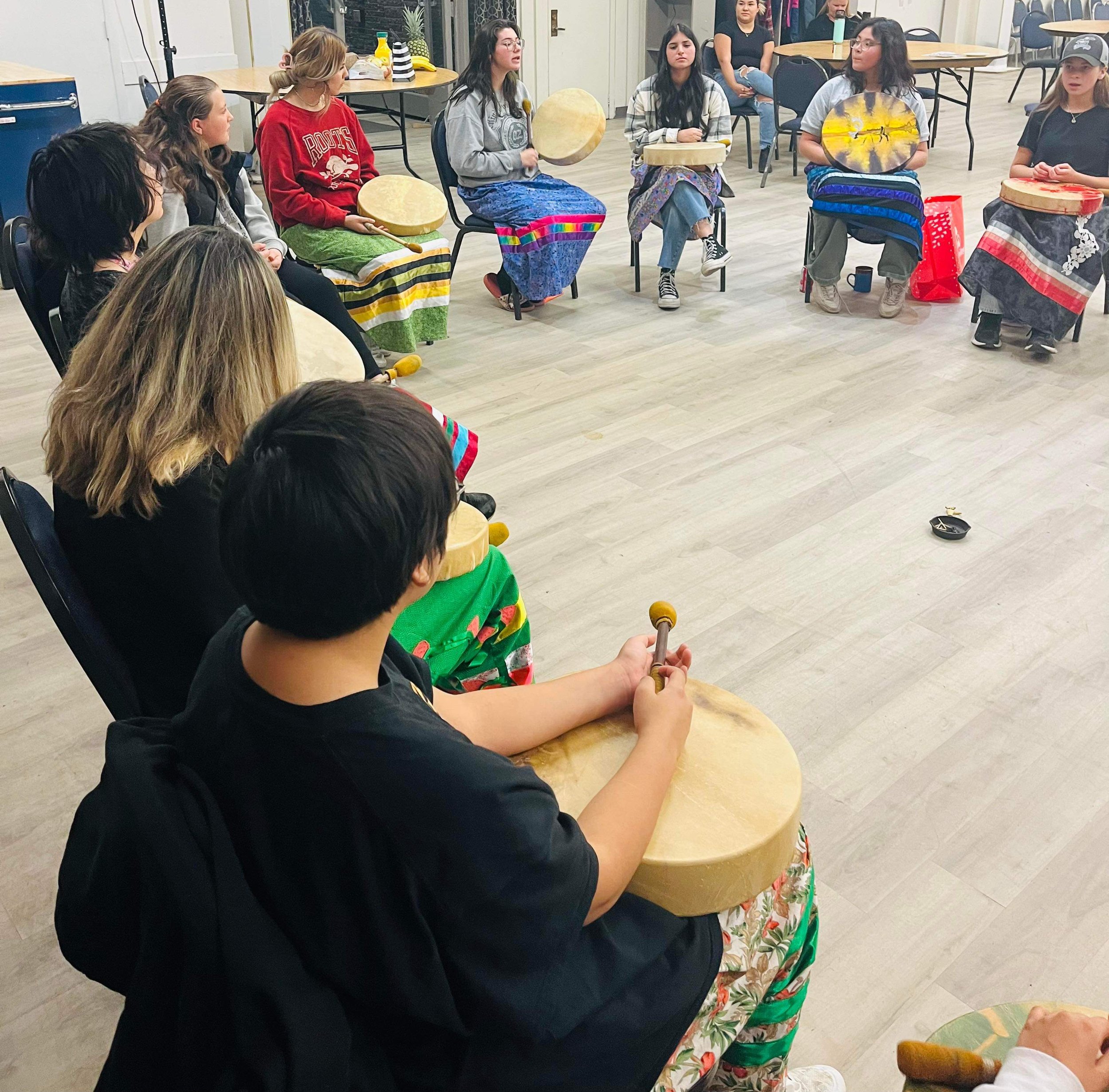 Group of children and teenagers sitting in a circle, playing drums indoors.