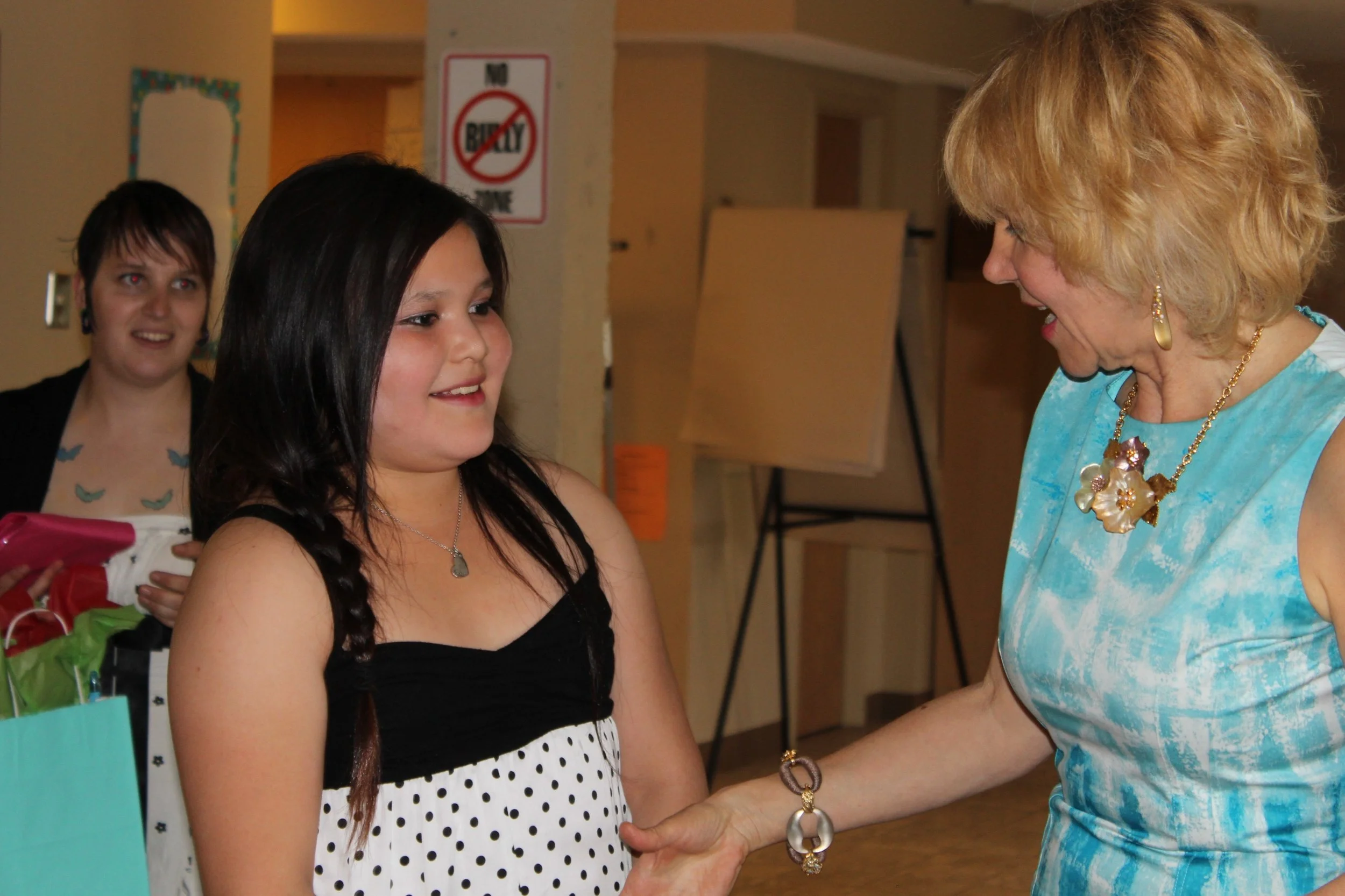 A young girl with long dark hair shaking hands with an older woman with blonde hair, both smiling, indoors with a sign that says 'No Bully Zone' in the background.