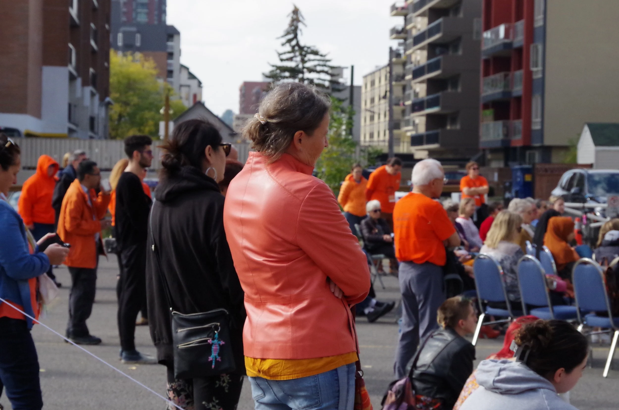 A diverse group of people, including women and children, gathered outdoors in an urban area, attending an event with chairs arranged in rows, some seated and others standing, with high-rise buildings and trees in the background.