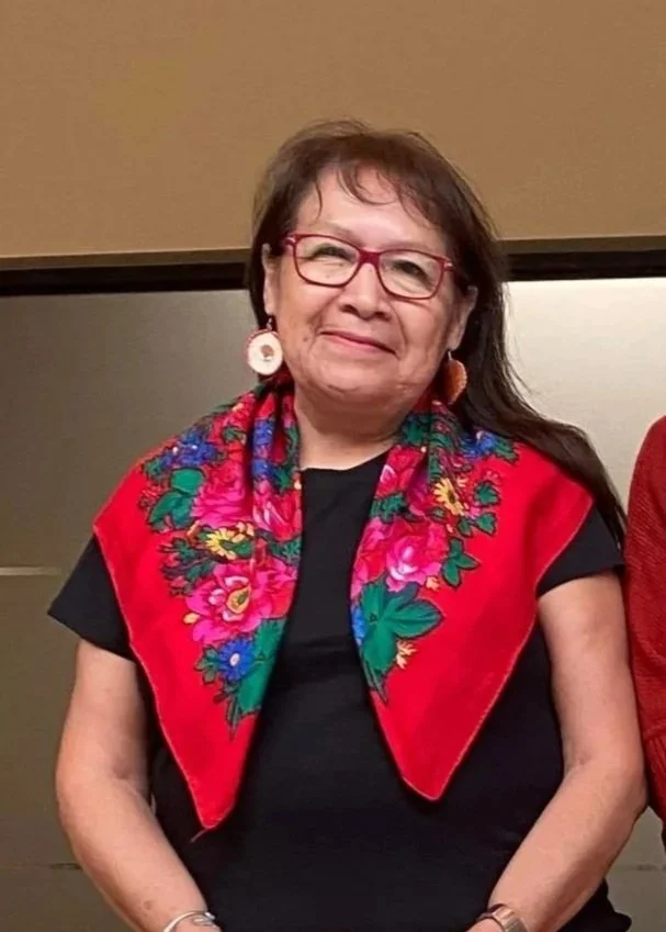 A woman with glasses and brown hair, wearing a red floral scarf and a black shirt, standing indoors.