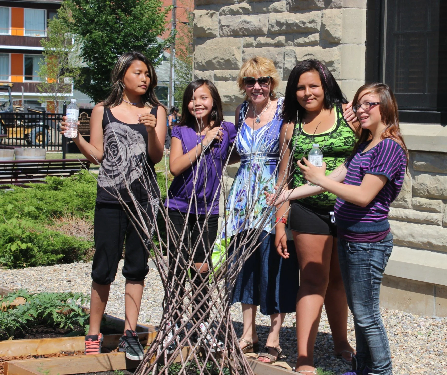 Group of five people, including four young girls and one adult woman, standing outdoors near a stone building with plants and trees in the background. They are gathered around a decorative lattice structure, with some holding water bottles and smilin