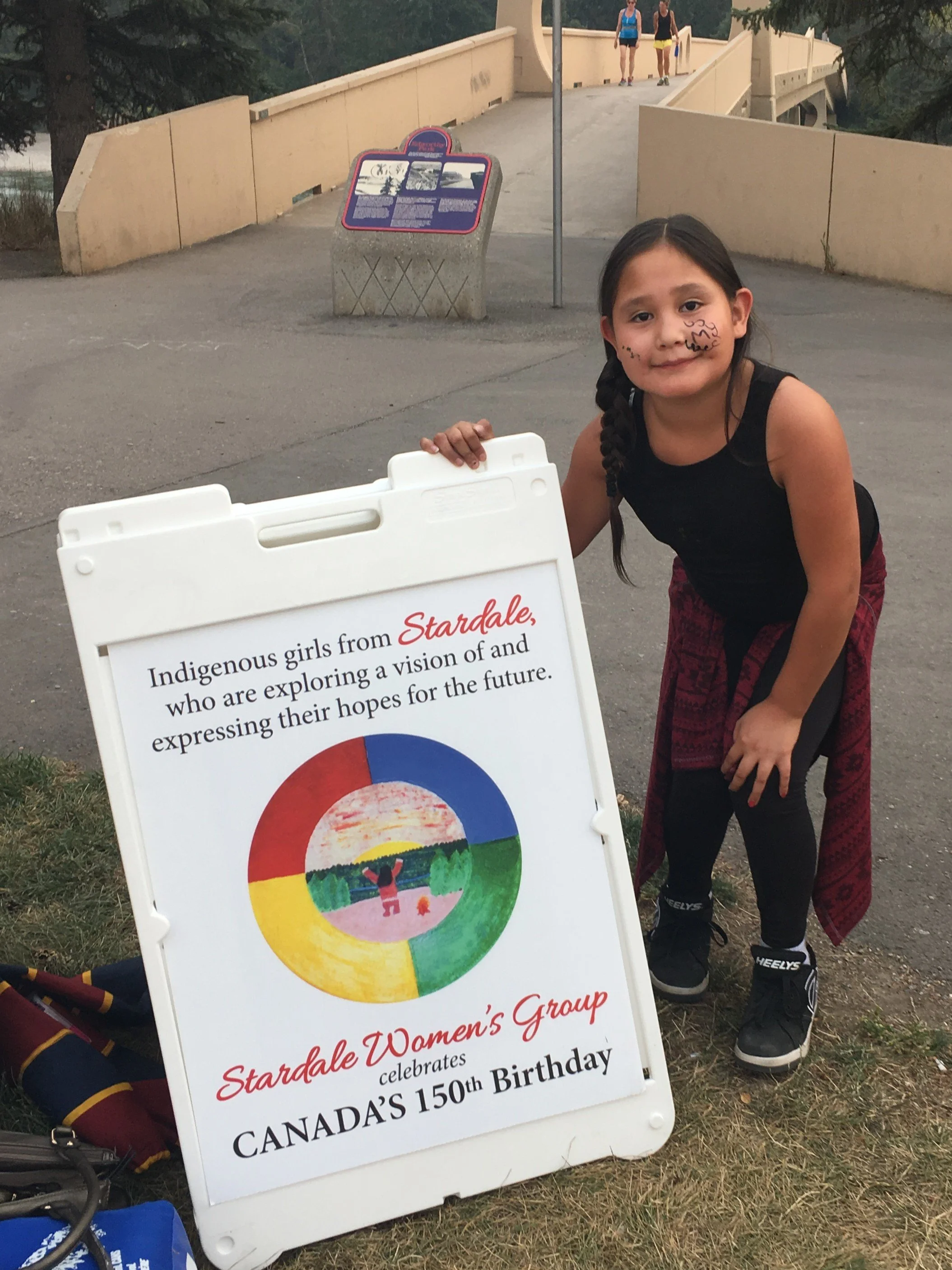 A young girl with face painting and a braid, wearing a black tank top and red patterned skirt, kneels next to an informational sign celebrating the 150th birthday of Canada, promoting Indigenous girls from Stardale expressing their hopes for the futu
