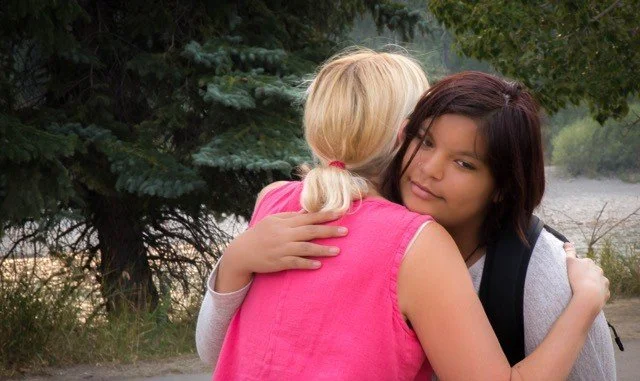 Two women hugging outdoors, one with blonde hair in a ponytail wearing a pink sleeveless top, and the other with dark hair, wearing a gray shirt and carrying a backpack, in front of trees and a body of water.