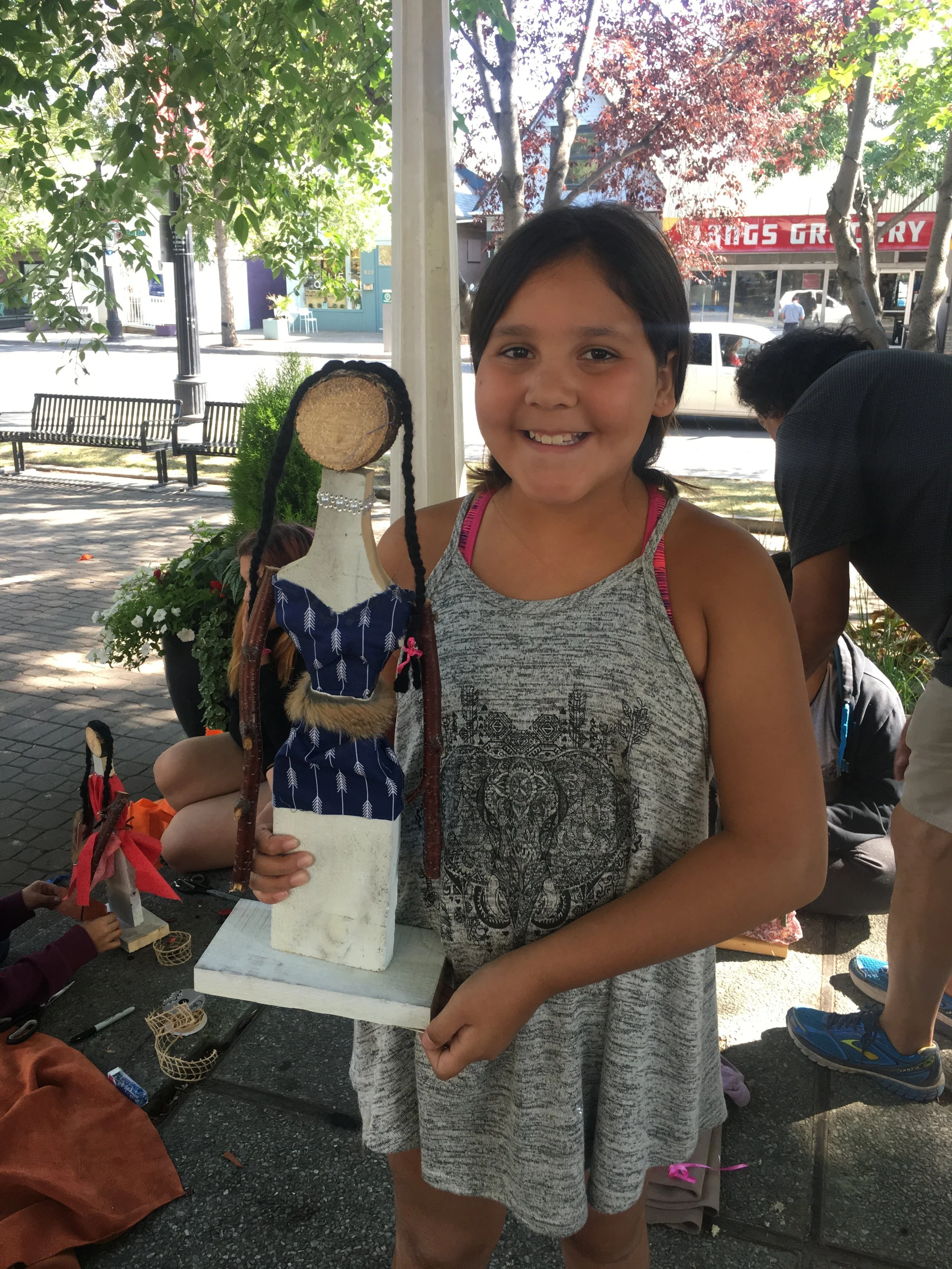 A young girl smiling and holding a handmade doll on a display stand outdoors in a park or street with trees, benches, and people in the background.