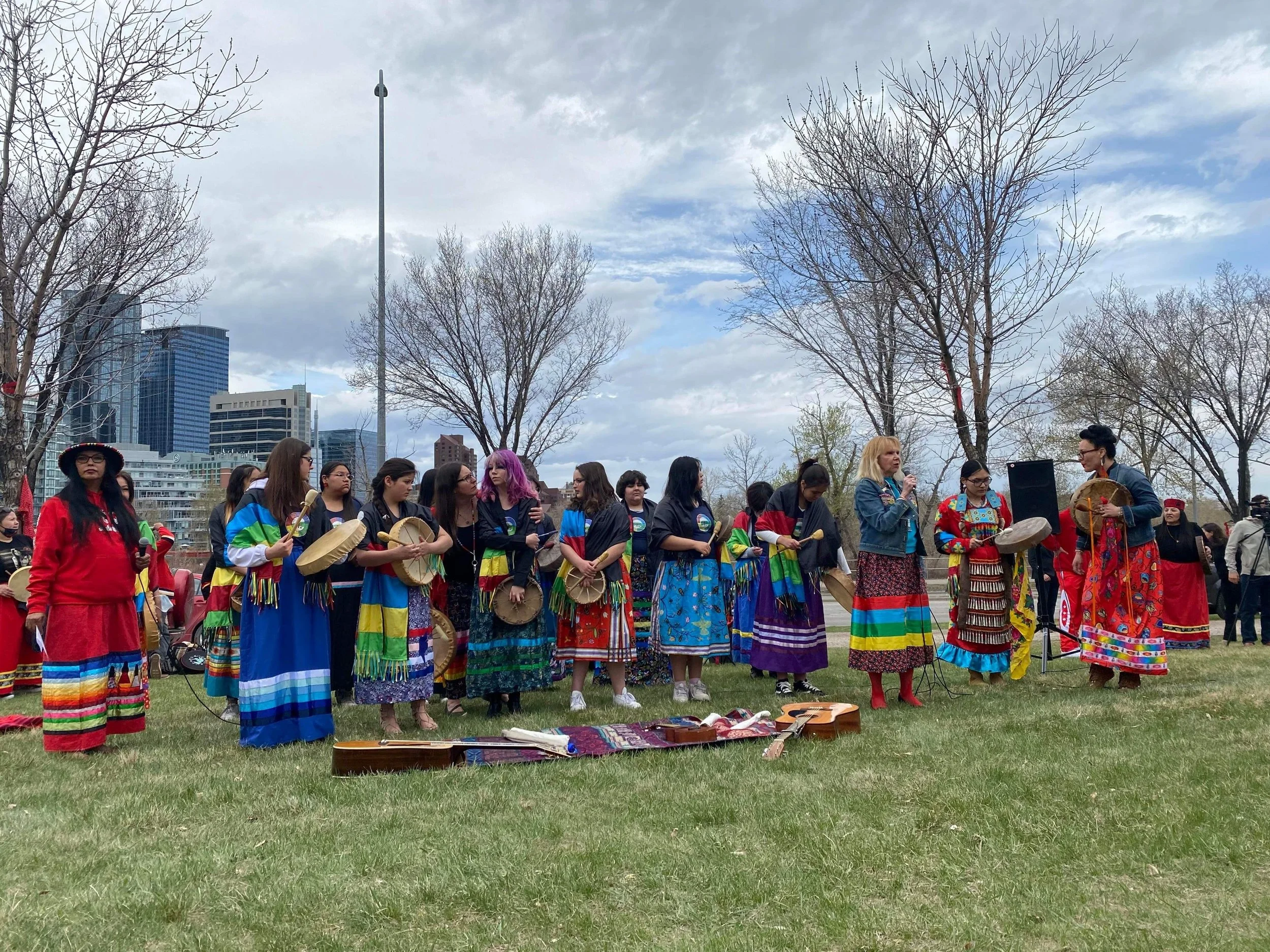Group of women dressed in colorful traditional clothing, standing outdoors on a grassy field with leafless trees and tall buildings in the background. Some women are holding drums, and one woman is speaking into a microphone.