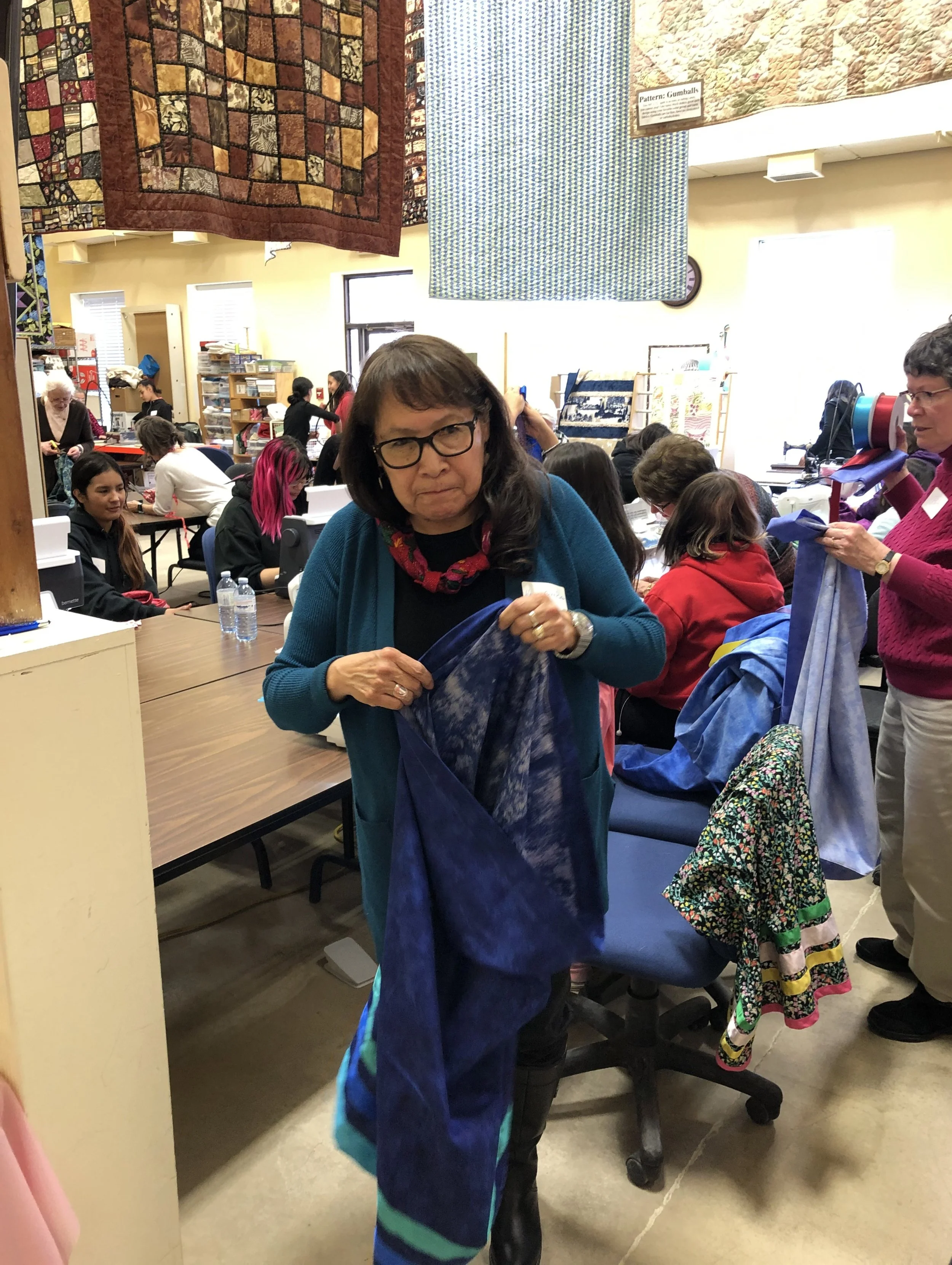 A woman with glasses and a blue cardigan holding up a piece of fabric in a quilt-making workshop with people working on Quilts and hanging fabrics in the background.