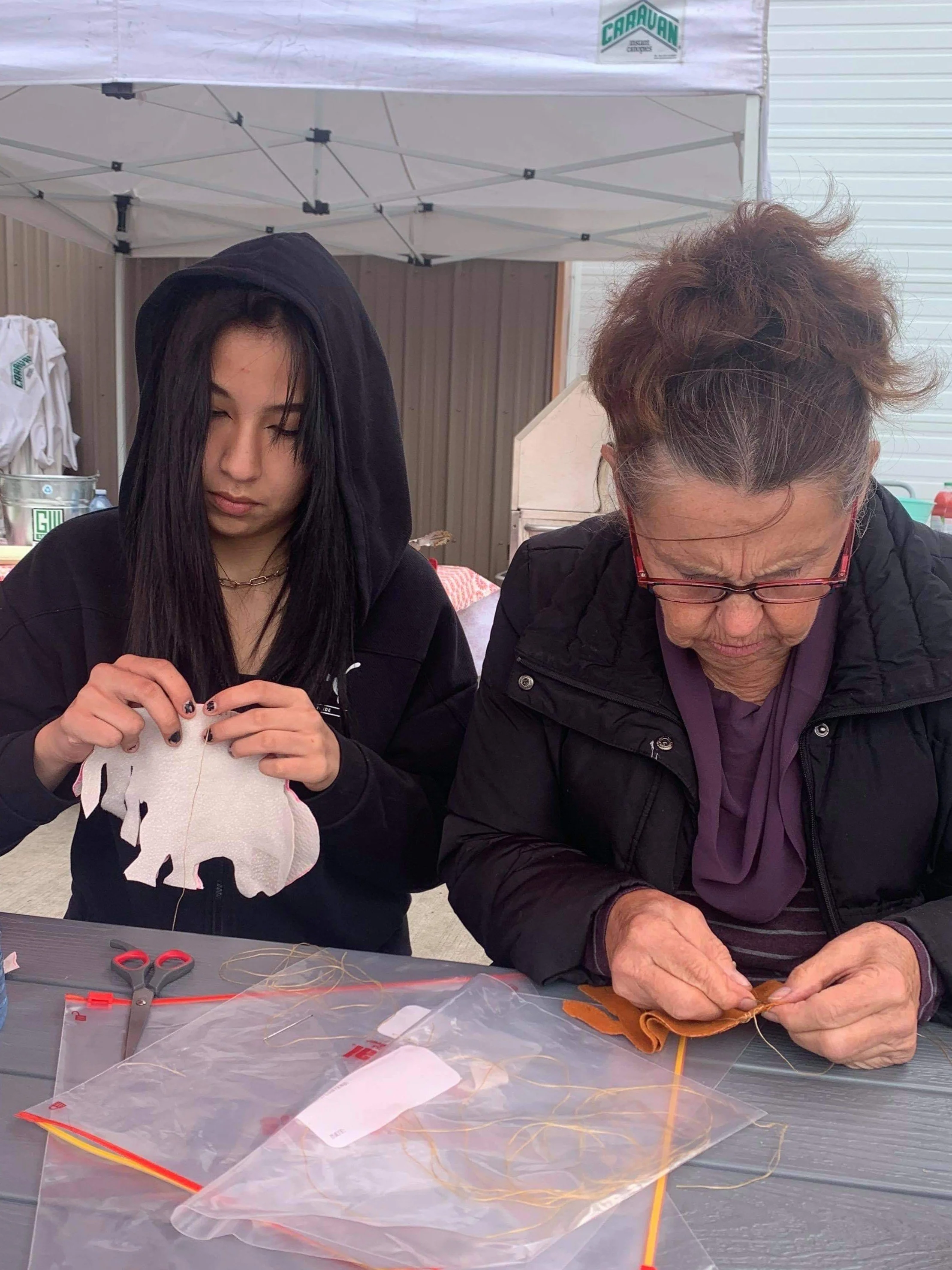 A young woman and an older woman are sitting at a table under a white canopy tent, focused on sewing projects. The young woman is wearing a black hoodie with the hood up and has long dark hair, while the older woman has curly hair tied up, glasses, a