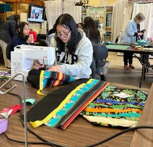 A woman using a sewing machine in a classroom, sewing colorful fabric with floral and striped patterns, while other students and instructors work in the background.