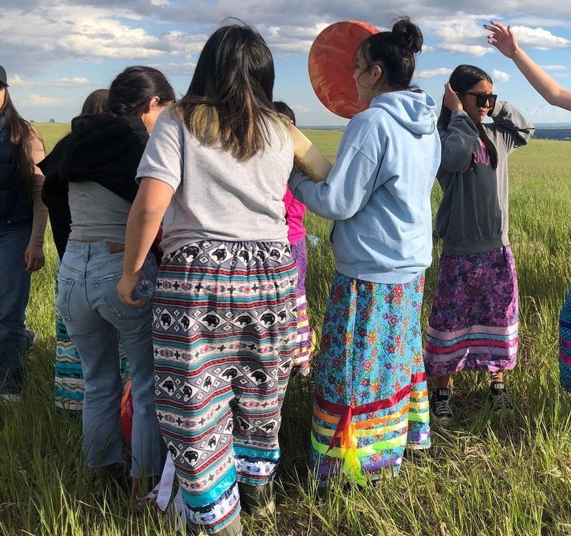 A group of people standing together in a grassy field, some wearing colorful skirts, with one person holding a red paper lantern, under a partly cloudy sky.
