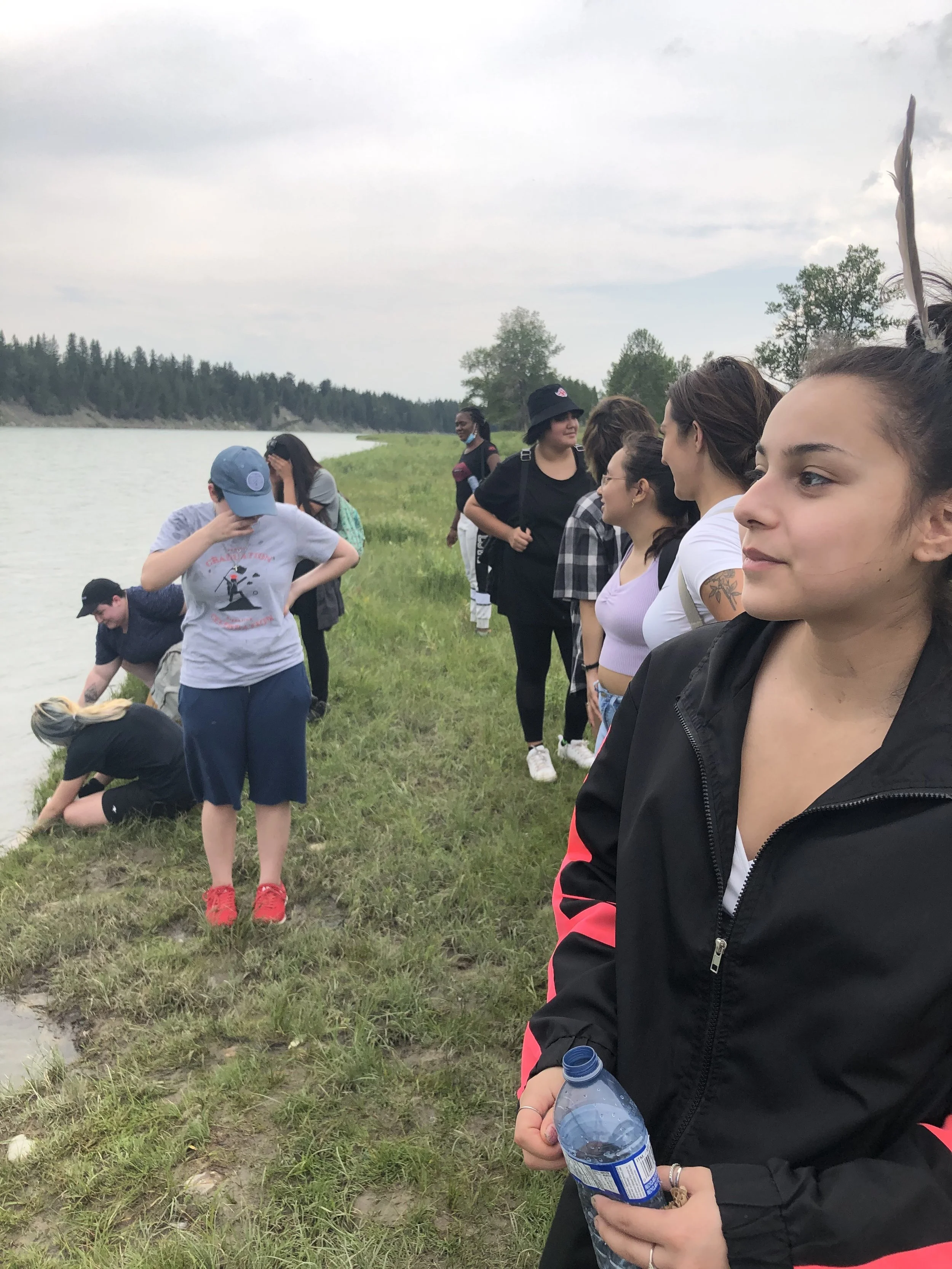 A group of people standing along a lakeshore on a cloudy day, some looking into the water and others observing the scenery.