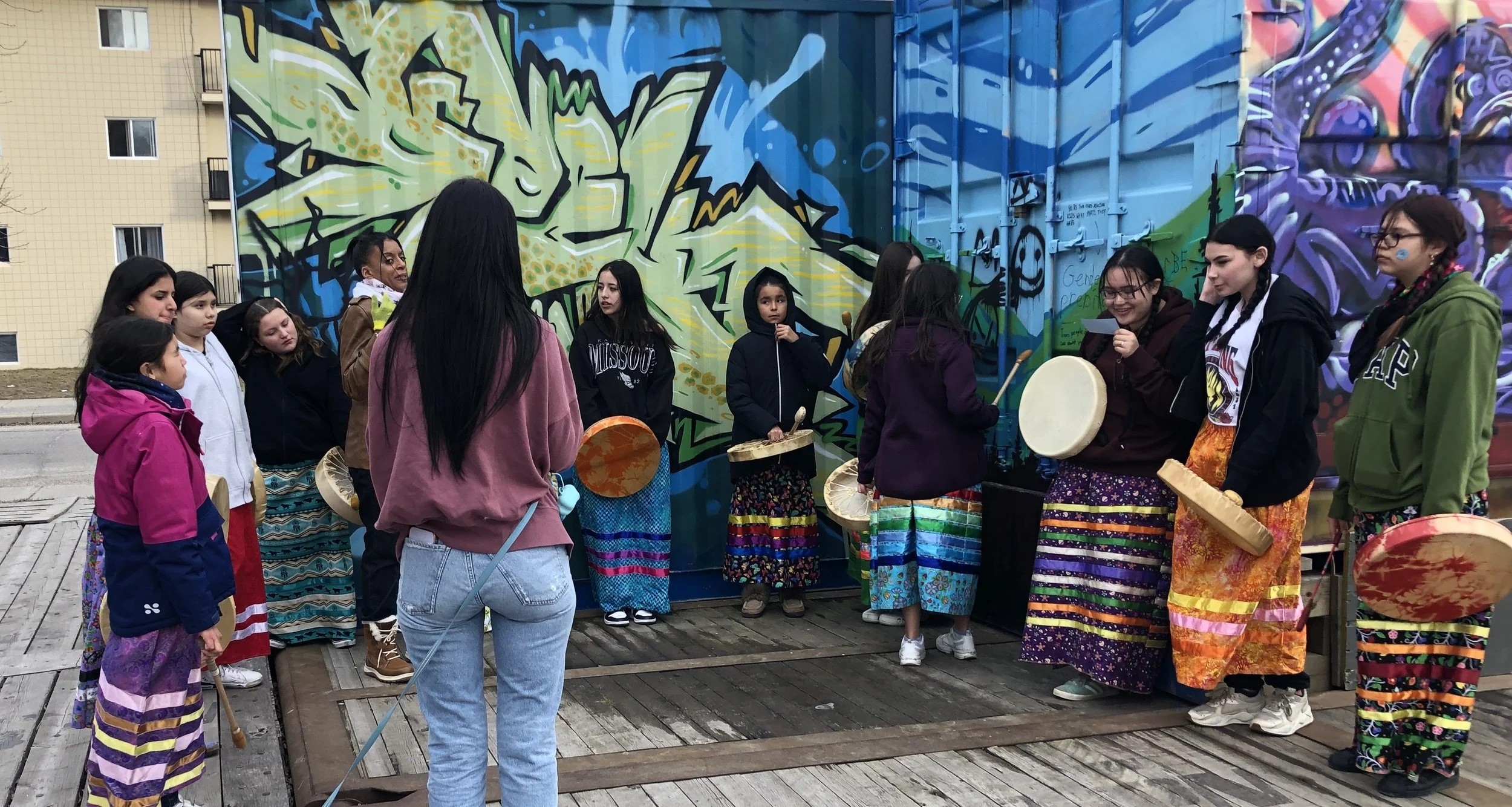 Group of young women and girls participating in a drum circle outdoors in front of a colorful graffiti mural, some holding drums and other percussion instruments.