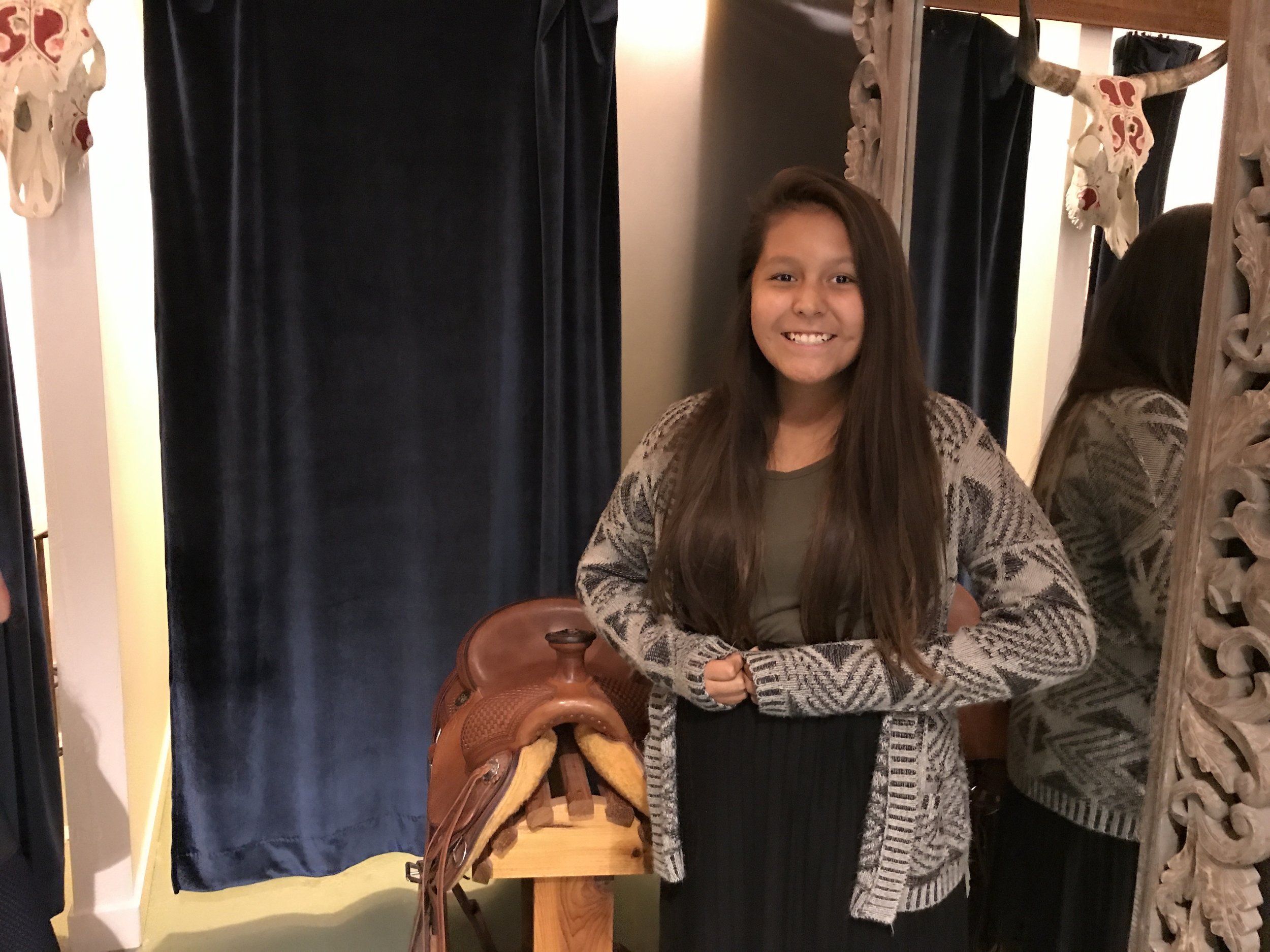 A young girl with long brown hair and a big smile, standing inside a fitting room or dressing area, wearing a gray T-shirt, a gray patterned cardigan, and a black skirt. She is standing next to a brown saddle on a wooden stand and mirror, with black 