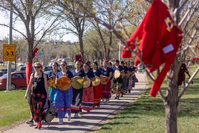 Group of women in traditional attire walking outdoors in a line, holding round decorative items, with trees and parked cars in the background, sunny day.