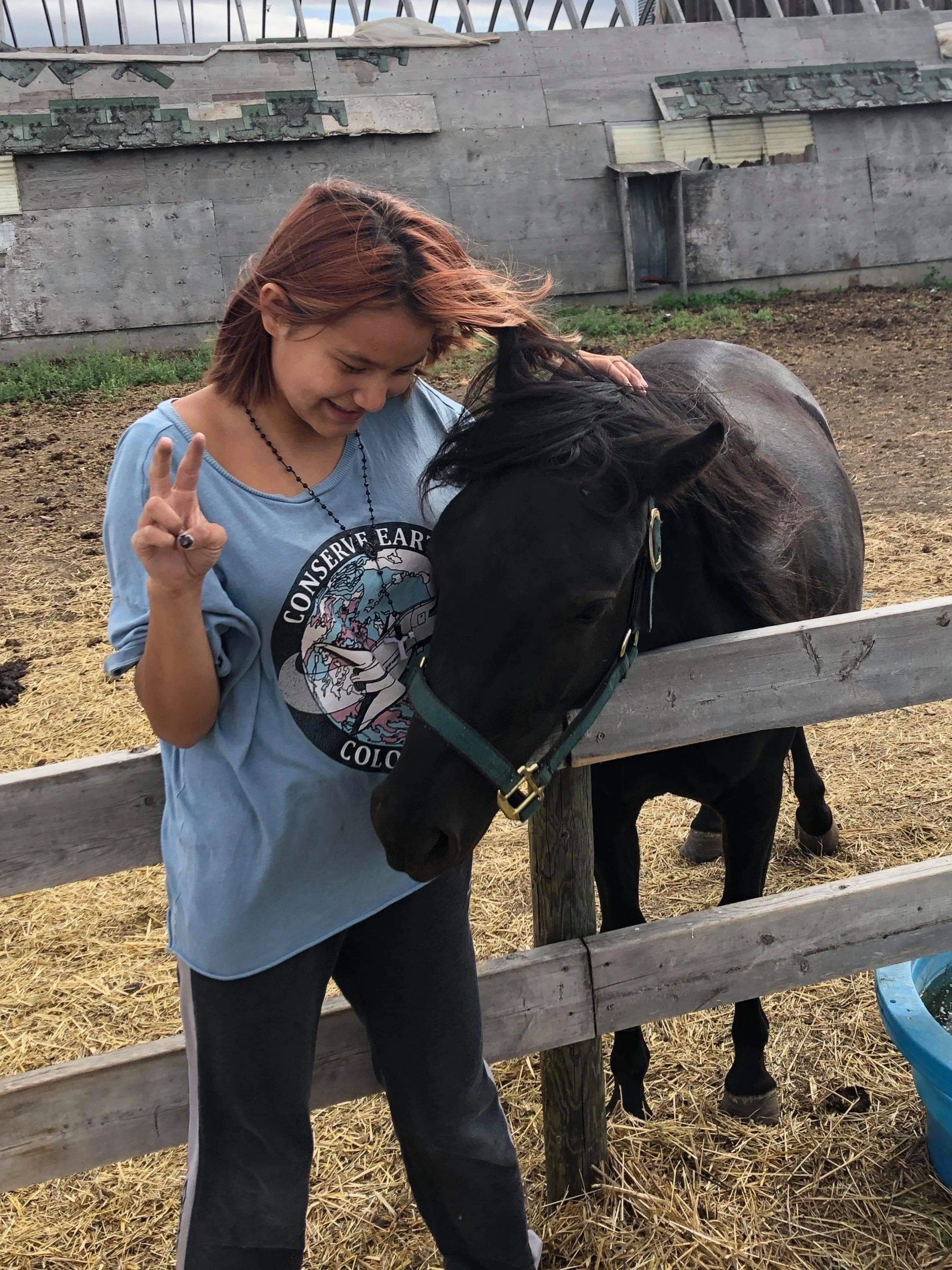 A woman with reddish hair and a blue t-shirt making a peace sign with her hand, smiling at a black horse behind a wooden fence.