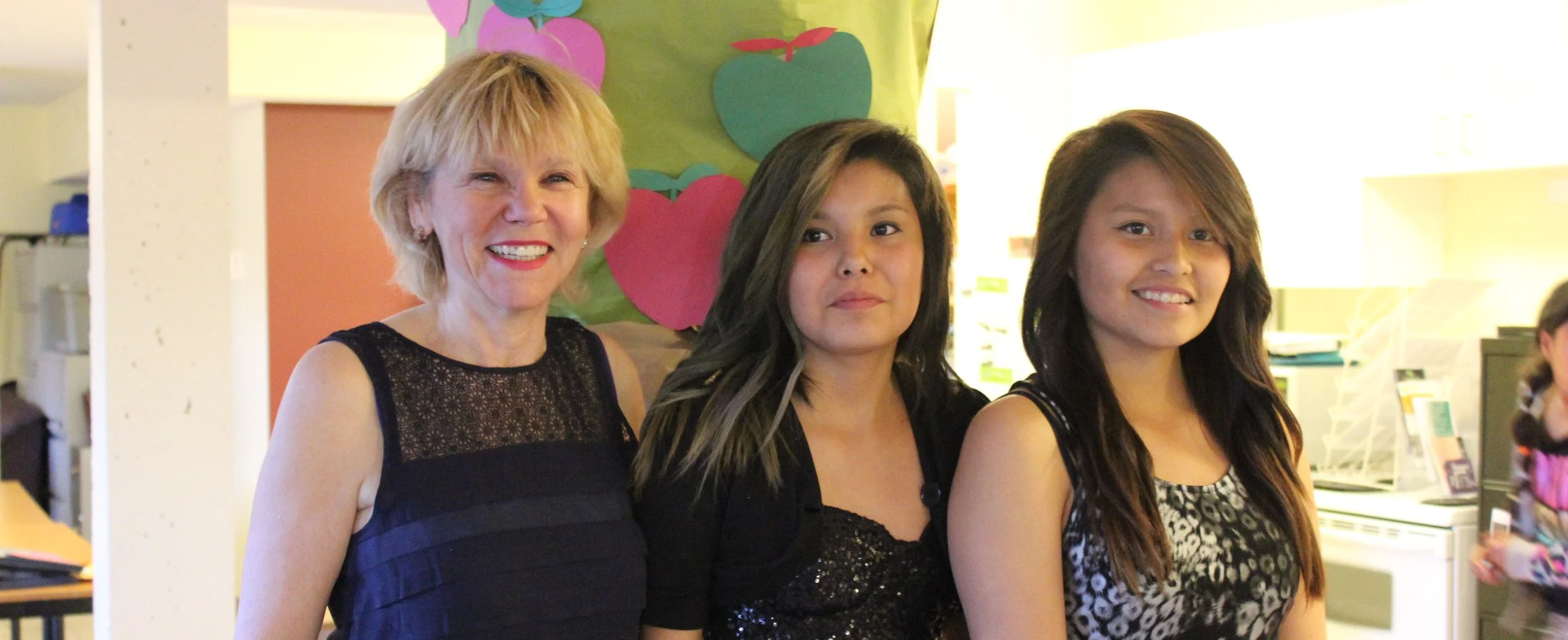 A group of three women standing together indoors. The woman on the left is older with short blonde hair, wearing a navy blue sleeveless top. The middle woman has long dark hair and is wearing a black top with a shiny pattern. The woman on the right h