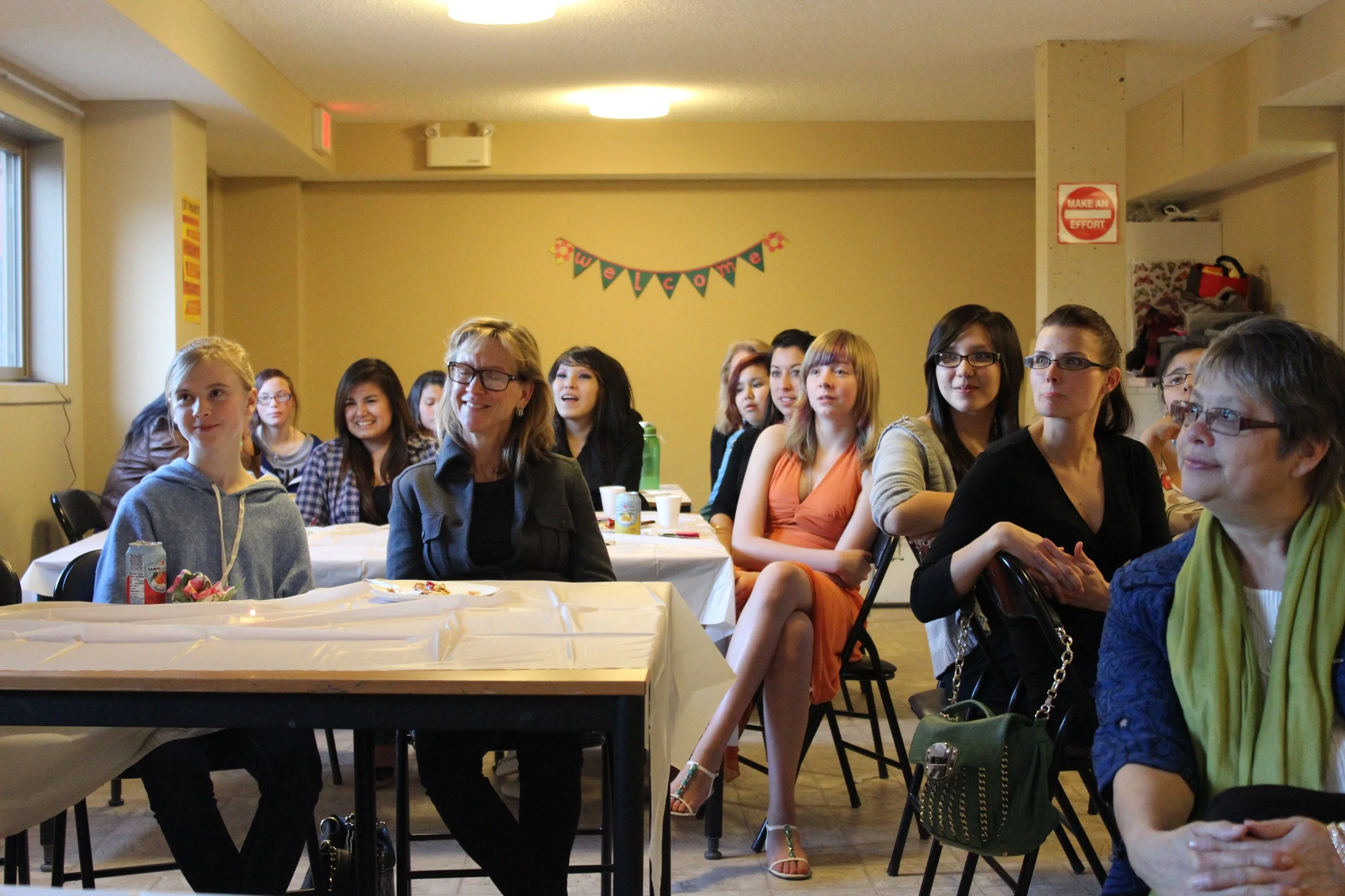 Group of women and girls sitting at a table in a room decorated with a "Welcome" banner, attending a gathering or celebration.