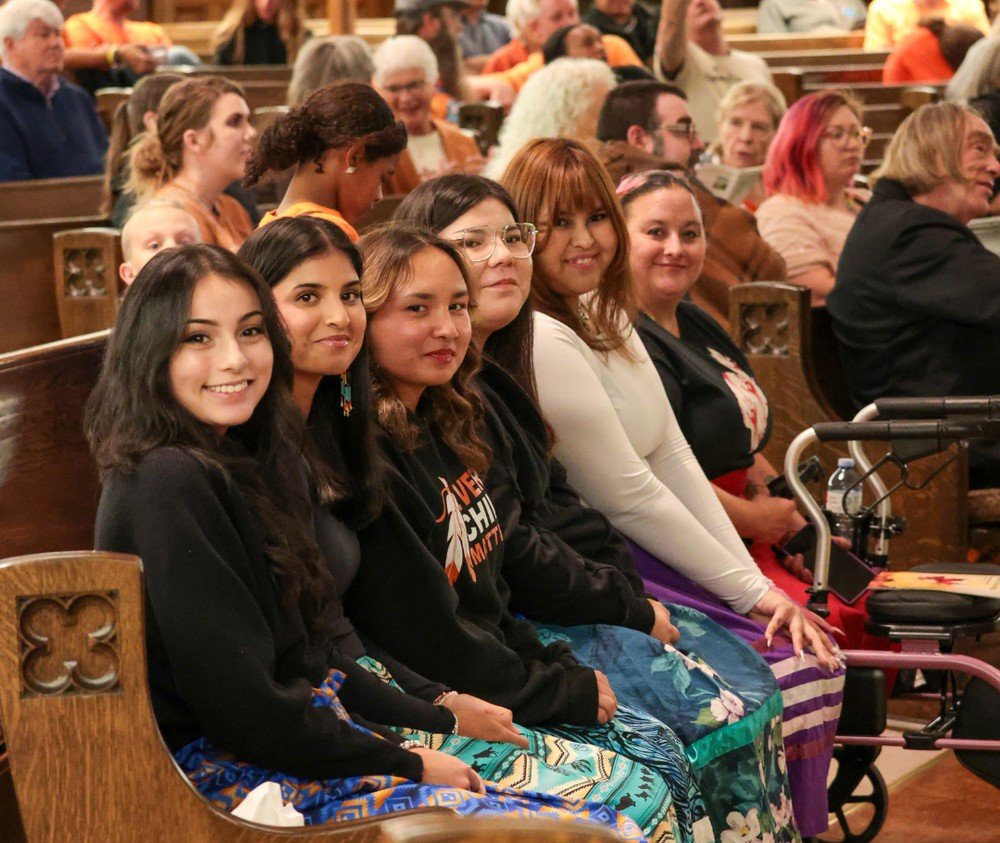 Group of women sitting in pews at an indoor event, smiling at the camera.