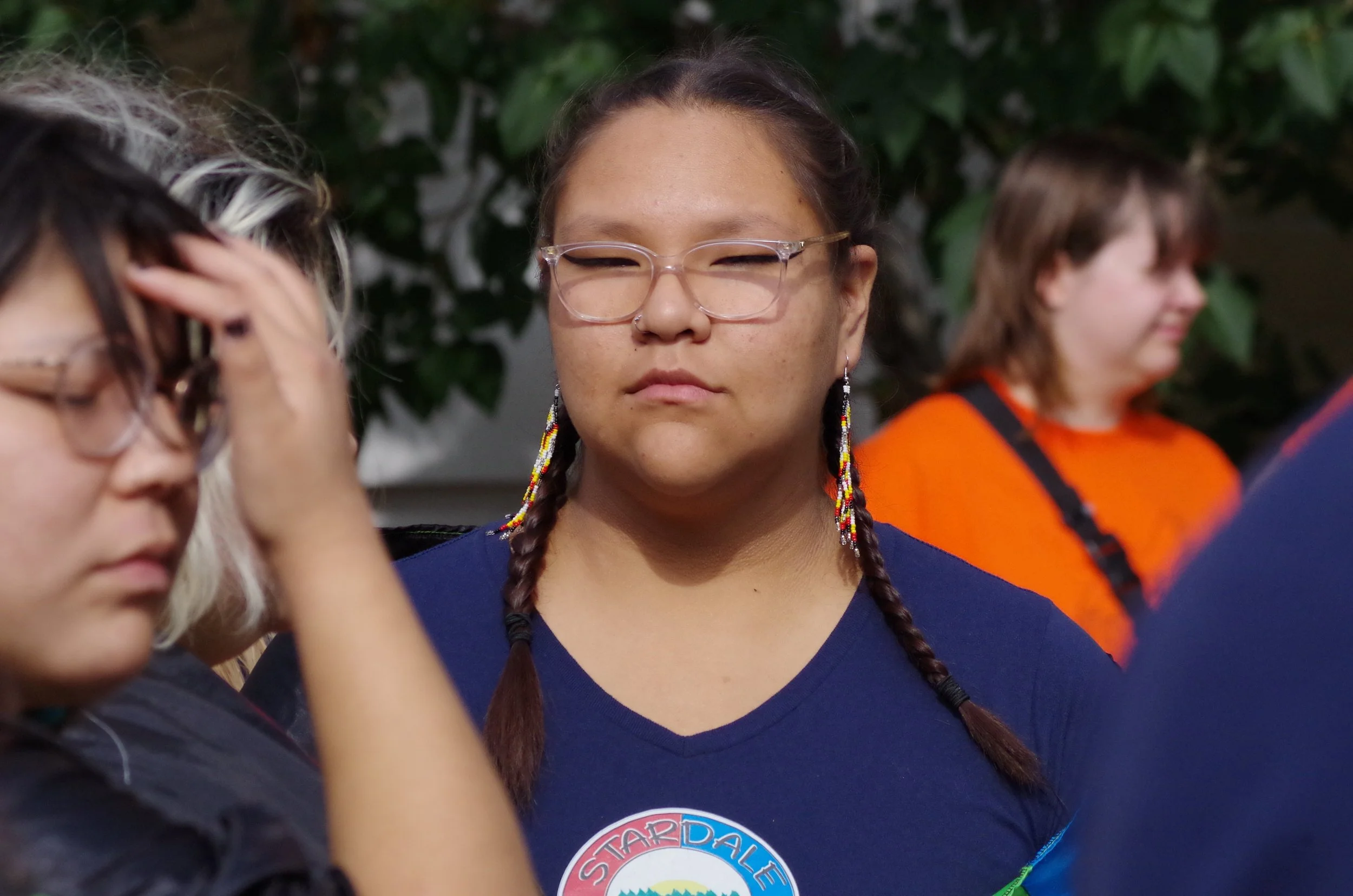 A woman with glasses, braided hair with beaded earrings, and a 'Stand for Dale' sticker on her shirt, appears contemplative or resting, with people around her, in an outdoor setting.