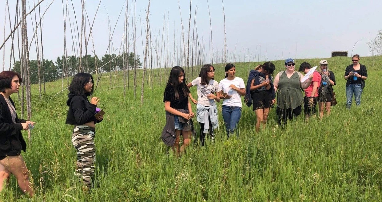 Group of women standing in a grassy field with tall, thin trees in the background, some holding notebooks and others reading or using their phones.