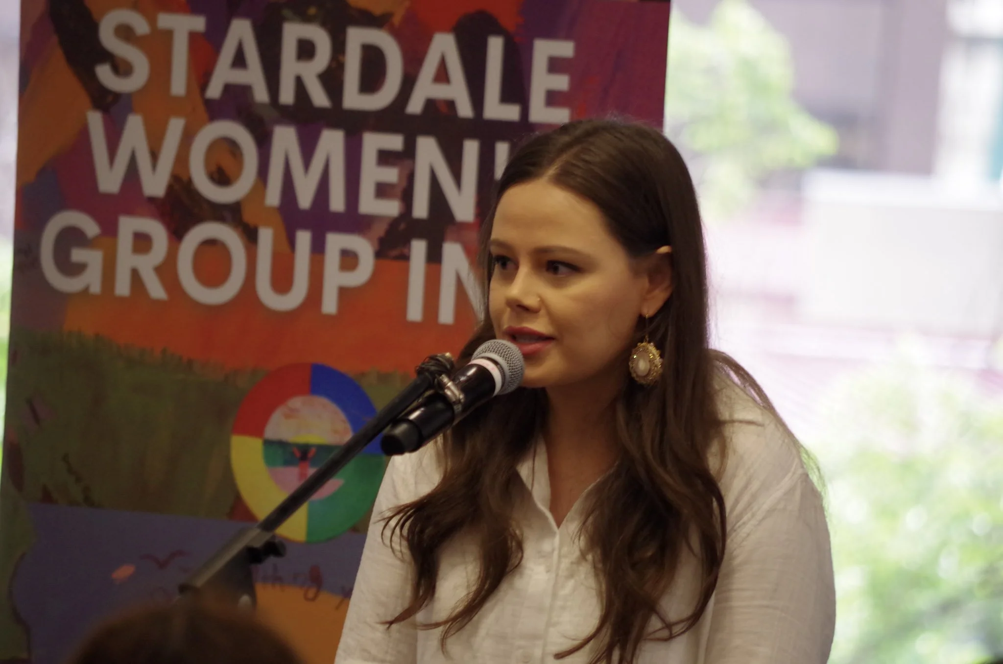 A woman with long brown hair and earrings speaking into a microphone at an event with a colorful banner behind her that reads 'Stardale Women's Group in'.