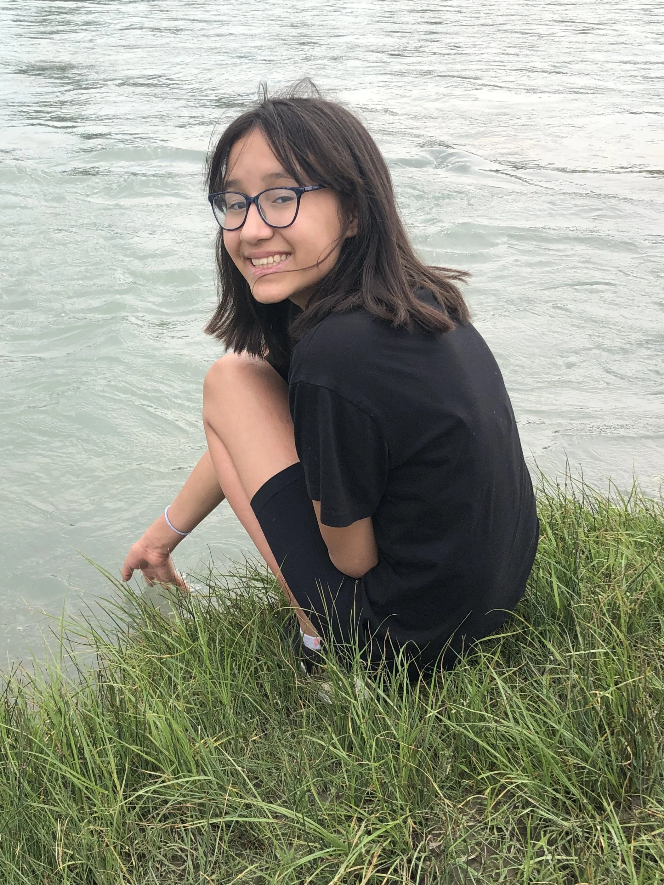 Young woman with glasses and black shirt sitting on grassy shoreline, dipping her hand into water.