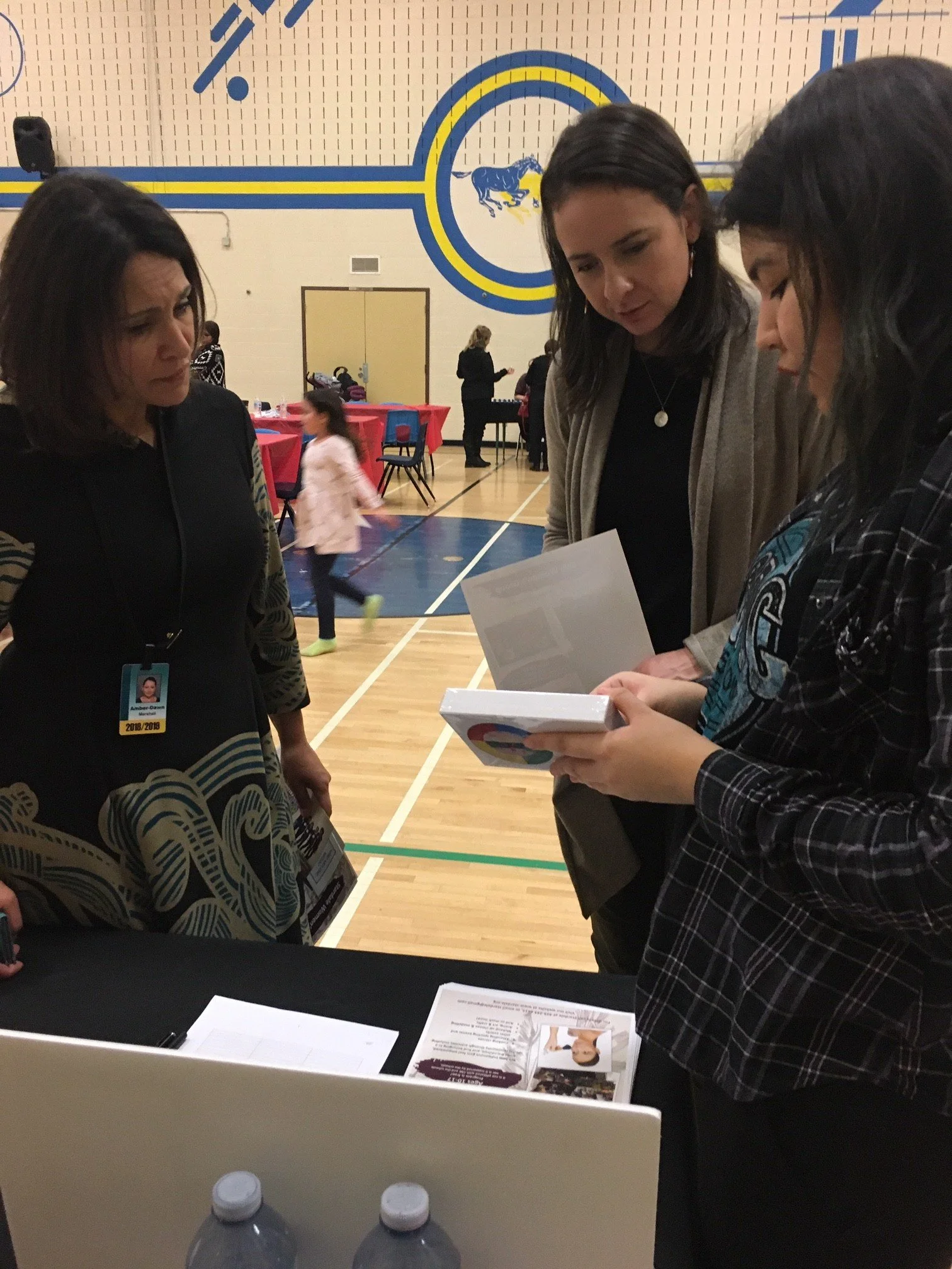 Three women are gathered around a table with papers and pamphlets in a gymnasium. One woman is holding a pamphlet, another is looking at a booklet, and the third appears to be listening. A few children are visible in the background, and there are tables with red tablecloths and chairs.