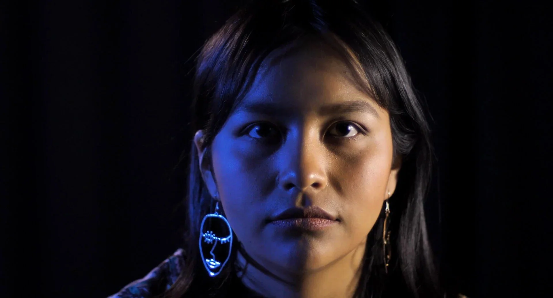Close-up of a woman with dark hair and earrings, illuminated with blue and neutral lighting on a black background.