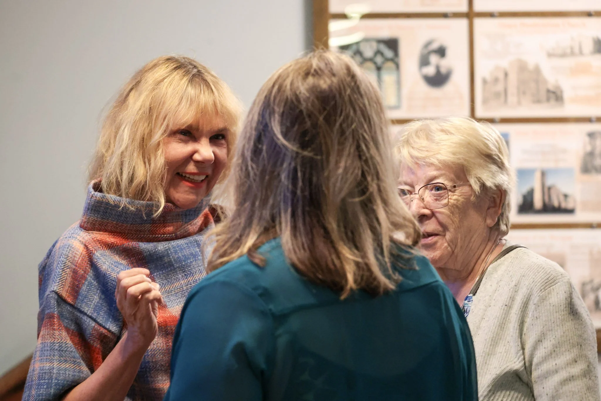 Three women having a conversation indoors, with photos and posters on the wall behind them.