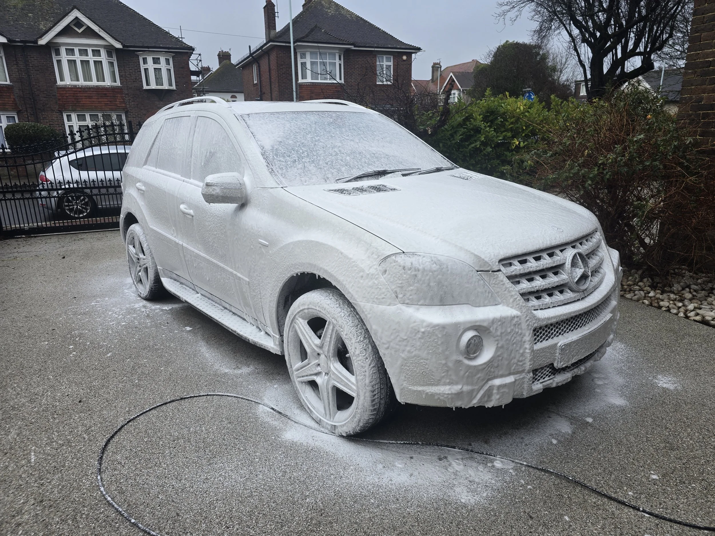 A Mercedes-Benz SUV covered in foam during a car wash on a residential driveway.