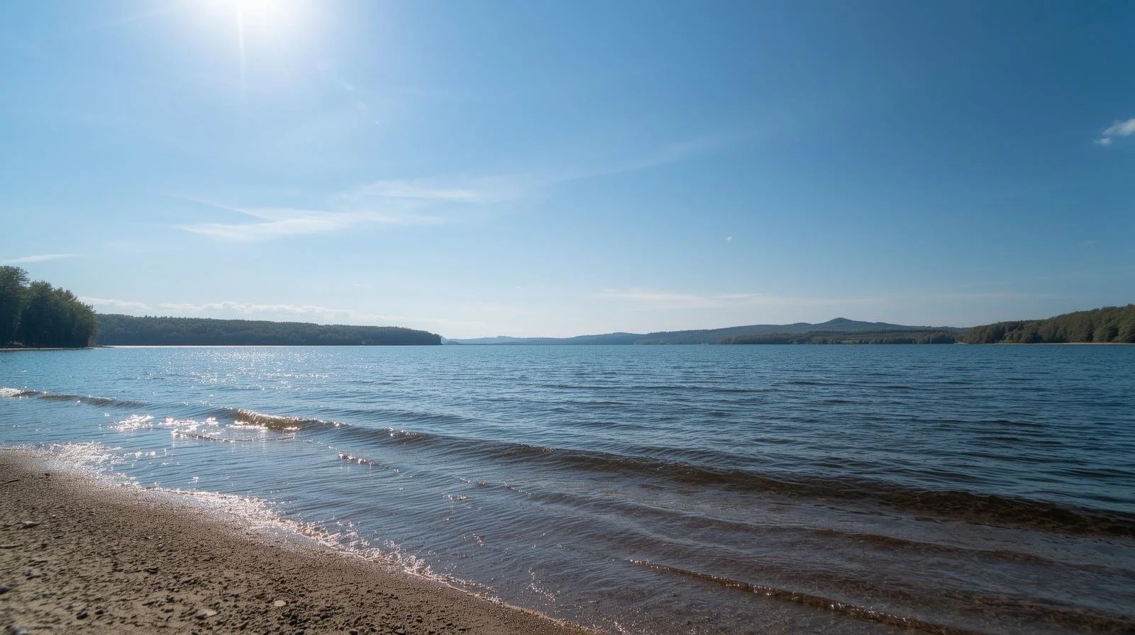 A peaceful lake with calm water and small waves near the sandy shoreline, surrounded by distant hills and a clear blue sky with bright sunlight.