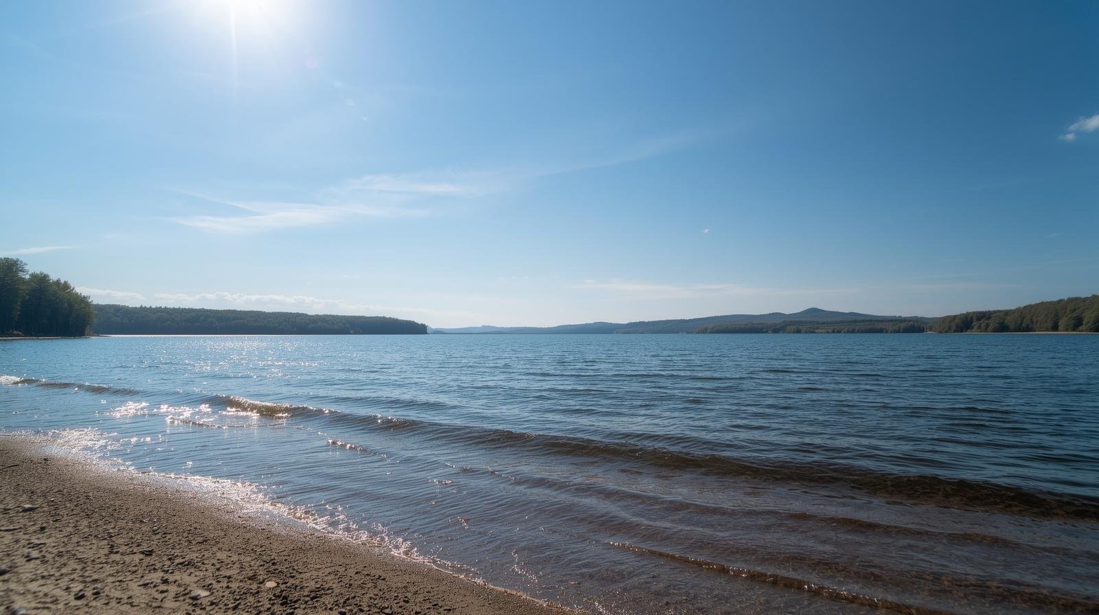 Calm lake with gentle waves near sandy shore, surrounded by trees and hills under a bright sunny sky.