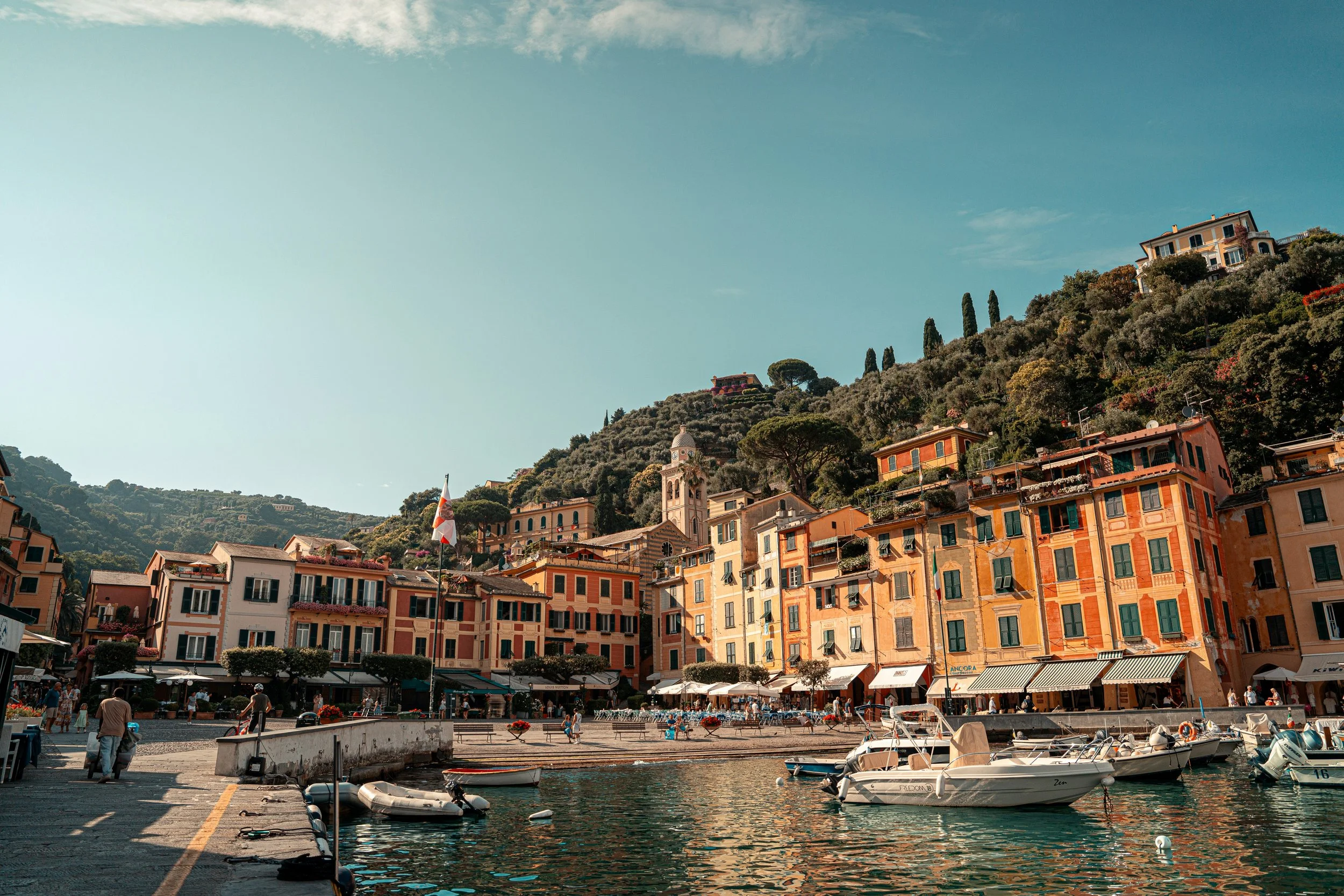 A coastal town with colorful buildings on a hillside overlooking a marina with boats, and a water front promenade filled with people, under a clear blue sky.