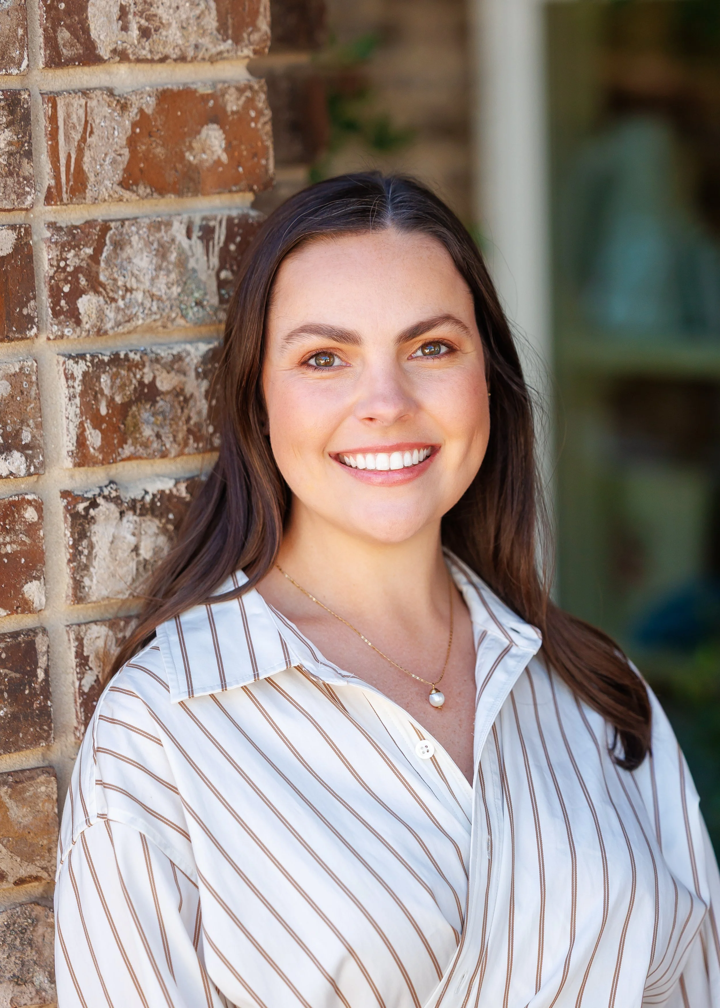 A woman with long brown hair, smiling, wearing a striped white and brown shirt, standing in front of a brick wall with greenery in the background.