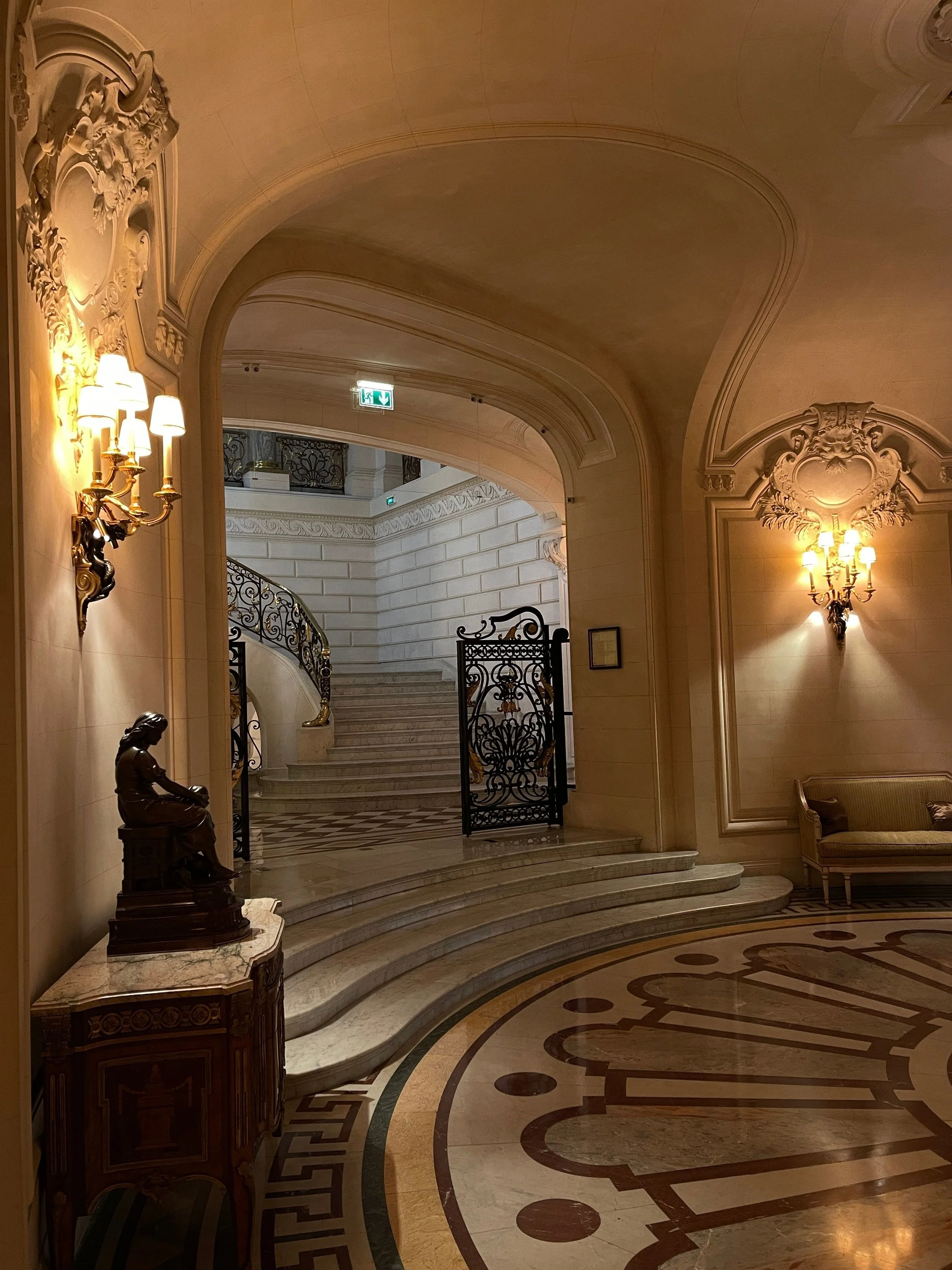 Luxurious interior with ornate wall sconces, marble staircase, intricate iron gate, marble bust statue on a wooden pedestal, marble flooring with intricate inlay, elegant chair, and detailed wall molding.