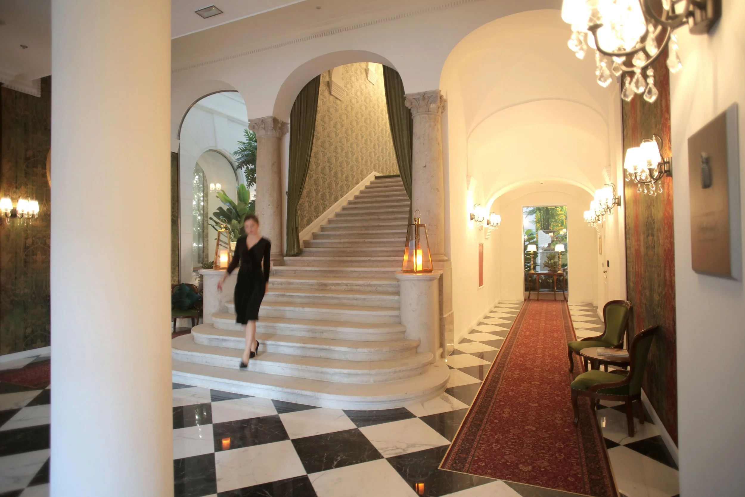 Elegant hotel lobby with a staircase framed by ornate columns, surrounded by warm lighting, chandeliers, and a patterned red carpet, with a blurred woman walking through.