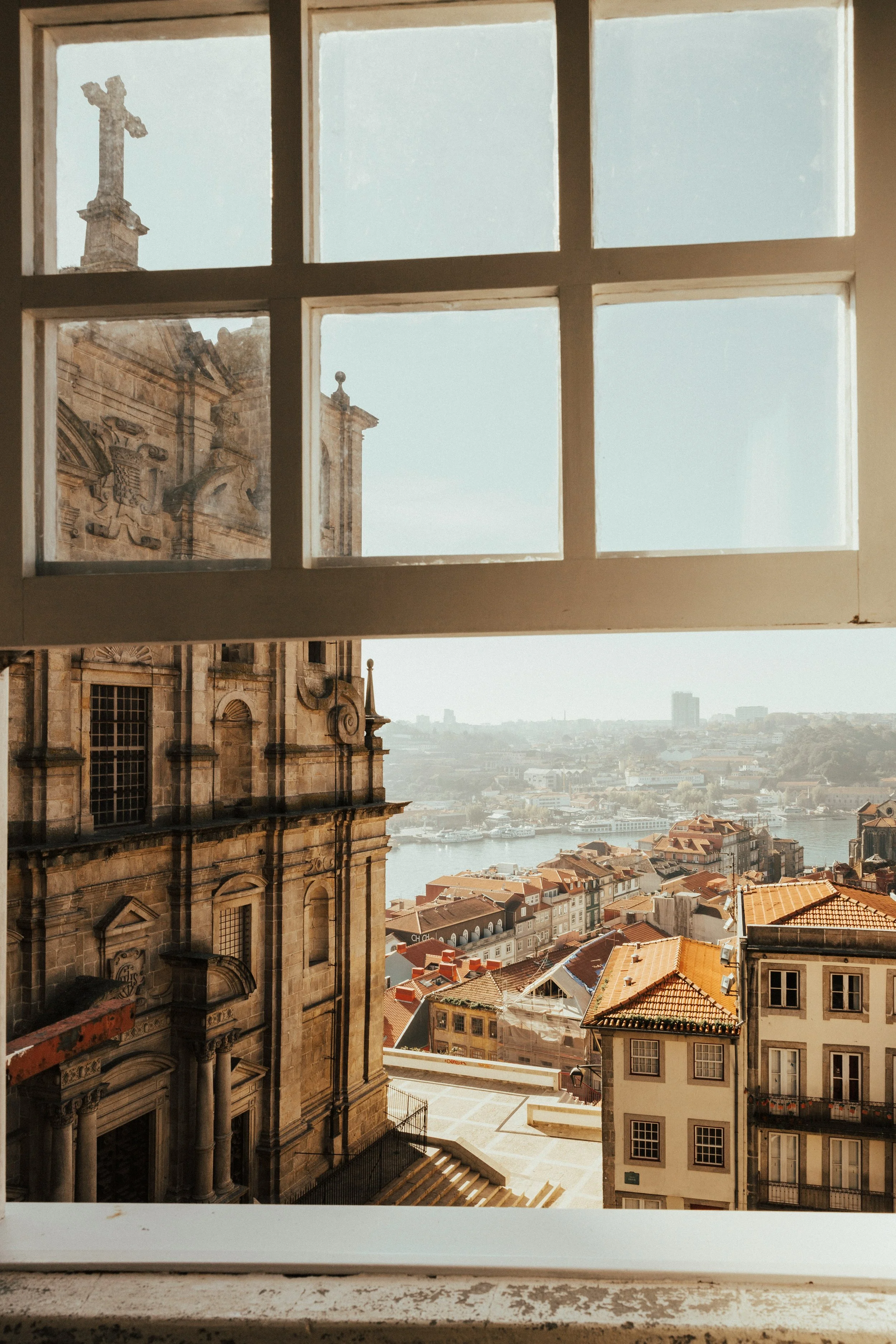 View from a window showing an old cathedral with ornate stonework and a cross on top, overlooking a cityscape with rooftops and a river in the distance.