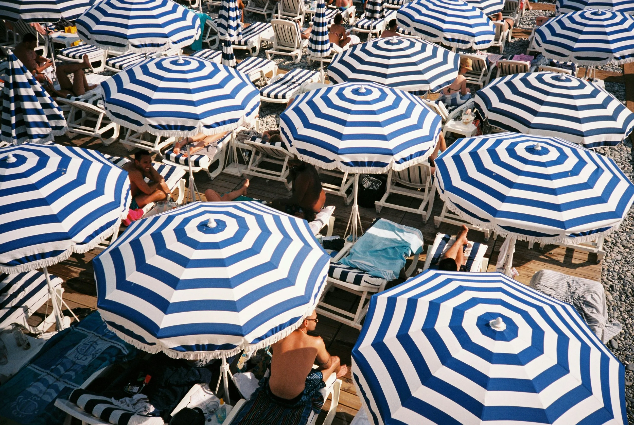 Beach scene with numerous people lounging on sunbeds under blue and white striped umbrellas on a pebbled shore.