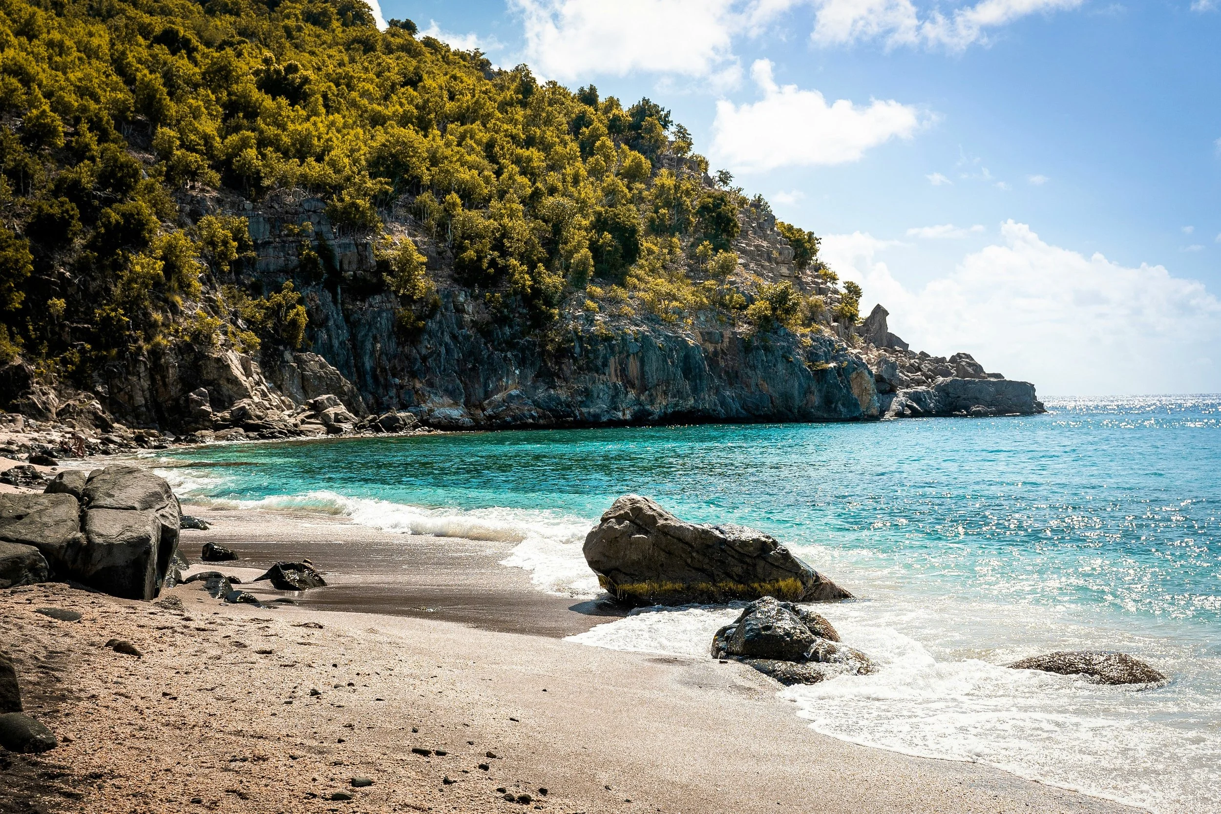 A scenic view of a beach with sandy shore and large rocks, green trees on a hillside, and blue ocean water under a partly cloudy sky.