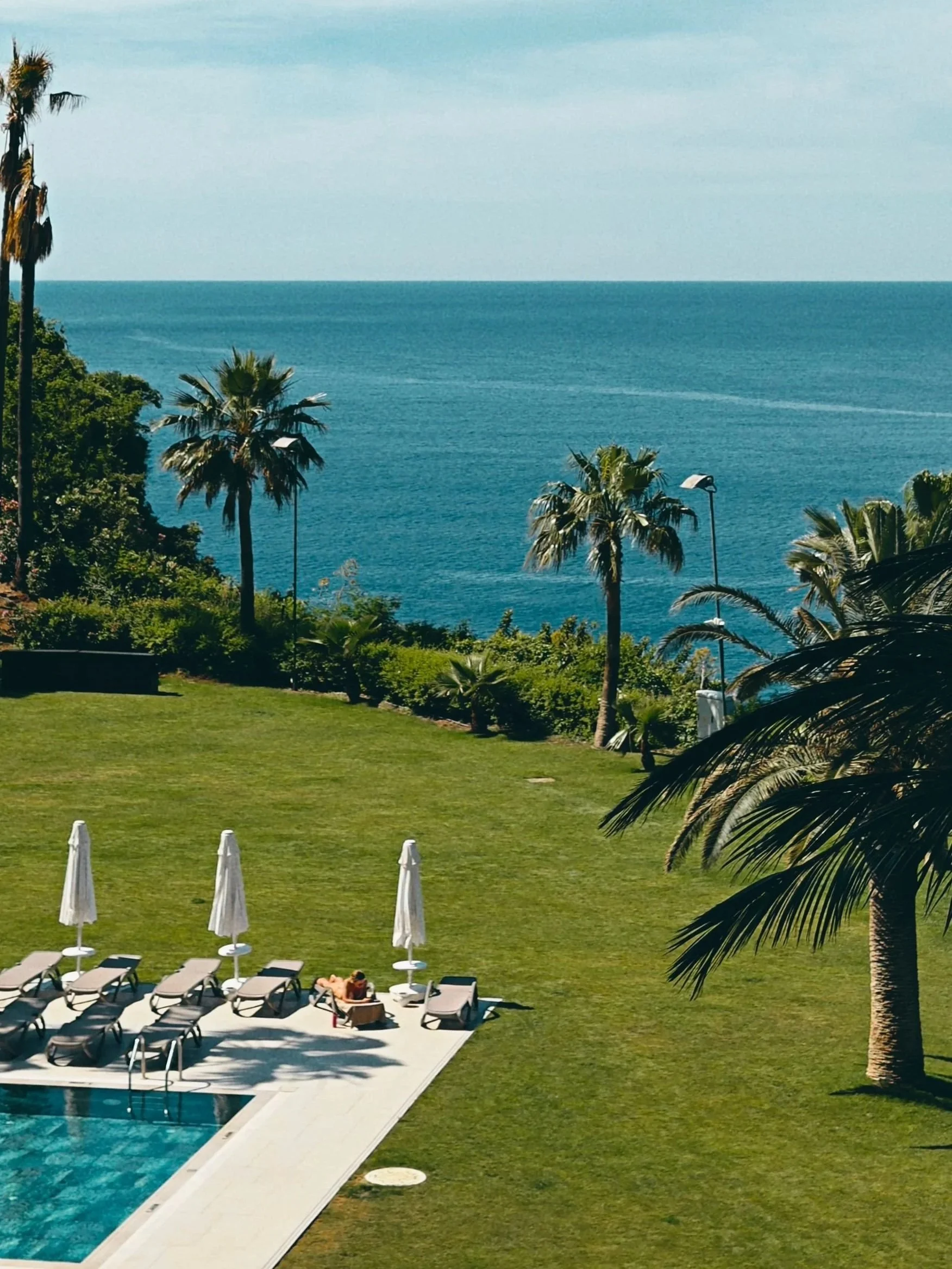 A swimming pool area with lounge chairs and umbrellas, tropical palm trees, and a view of the ocean in the background.
