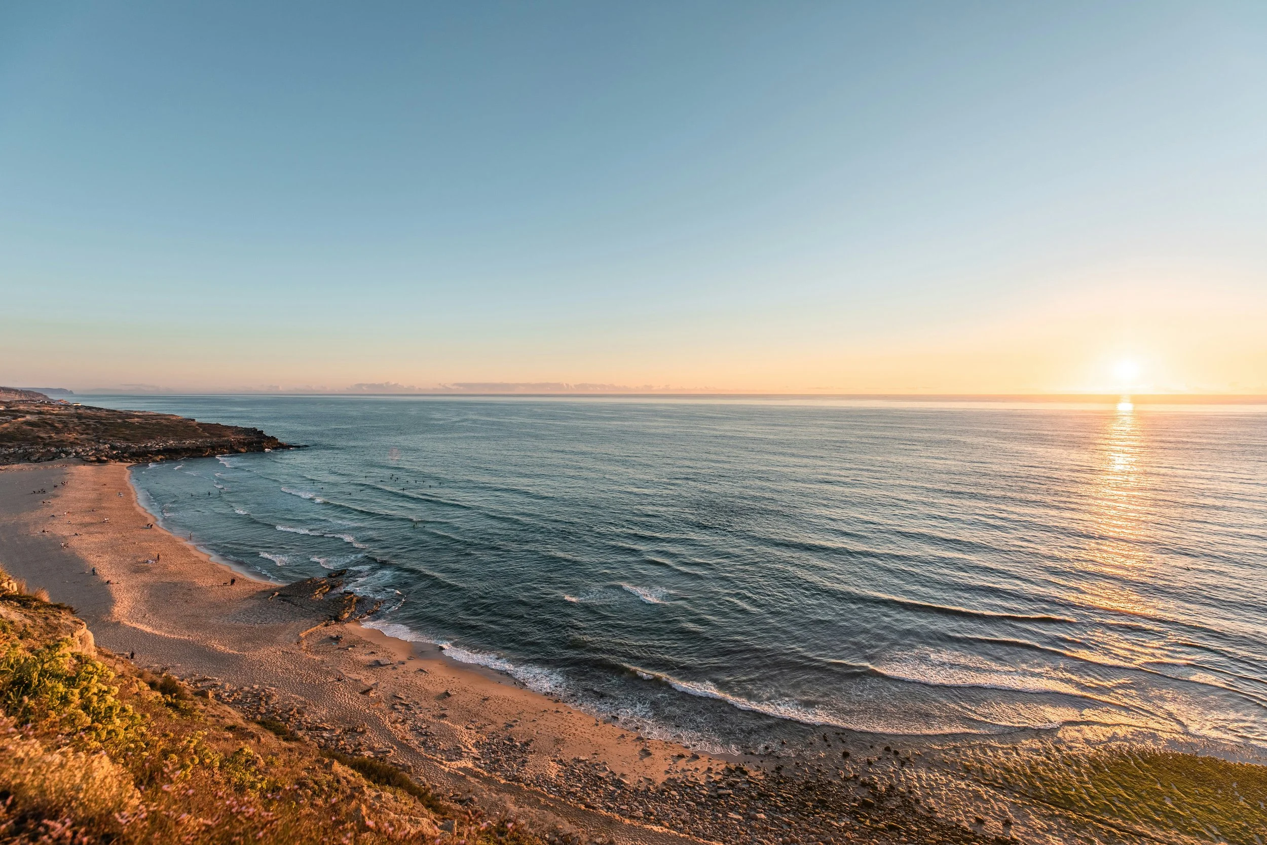 Coastal scene at sunset with sandy beach, waves, and cliffs on the left, and the sun low on the horizon over the ocean.
