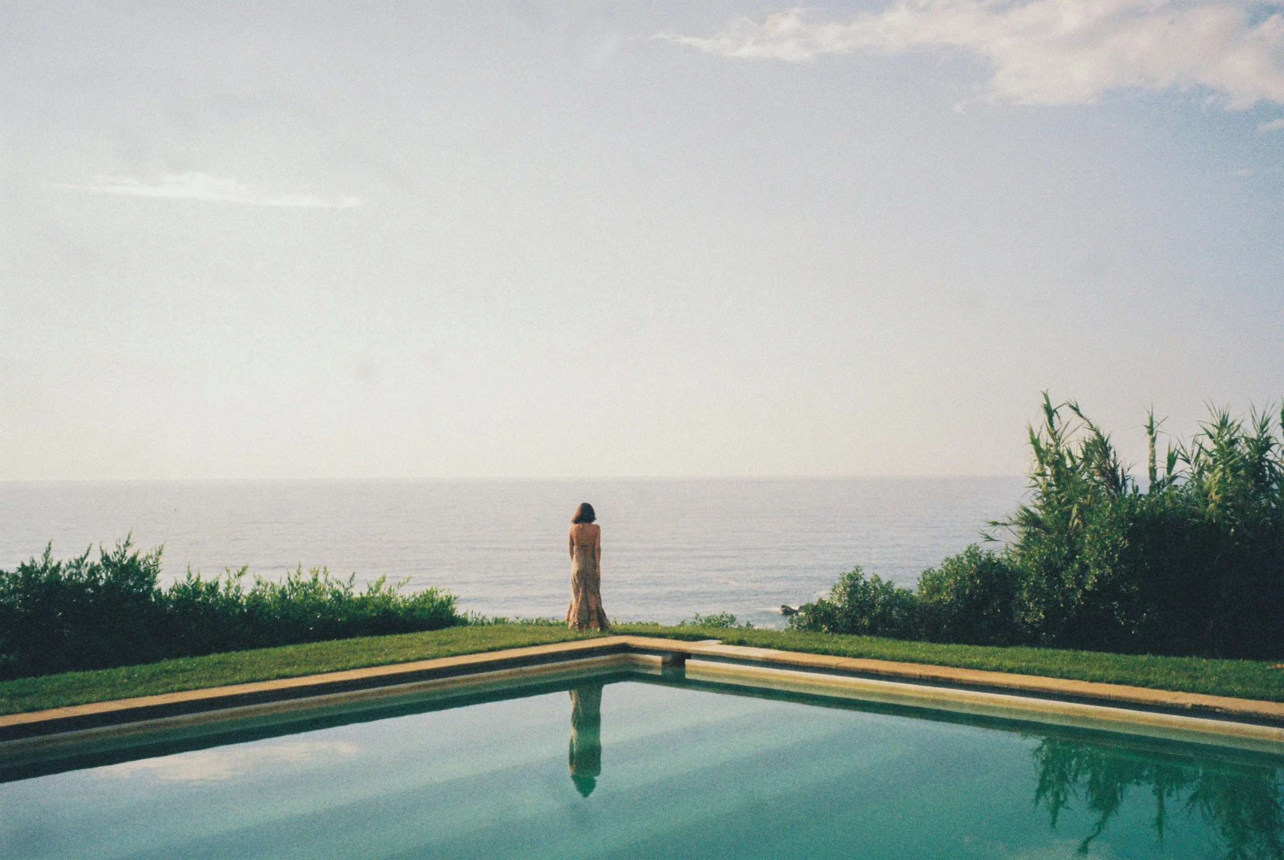 A woman in a beige dress standing by a pool, overlooking the ocean with greenery and bushes in the foreground, cloudy sky above.
