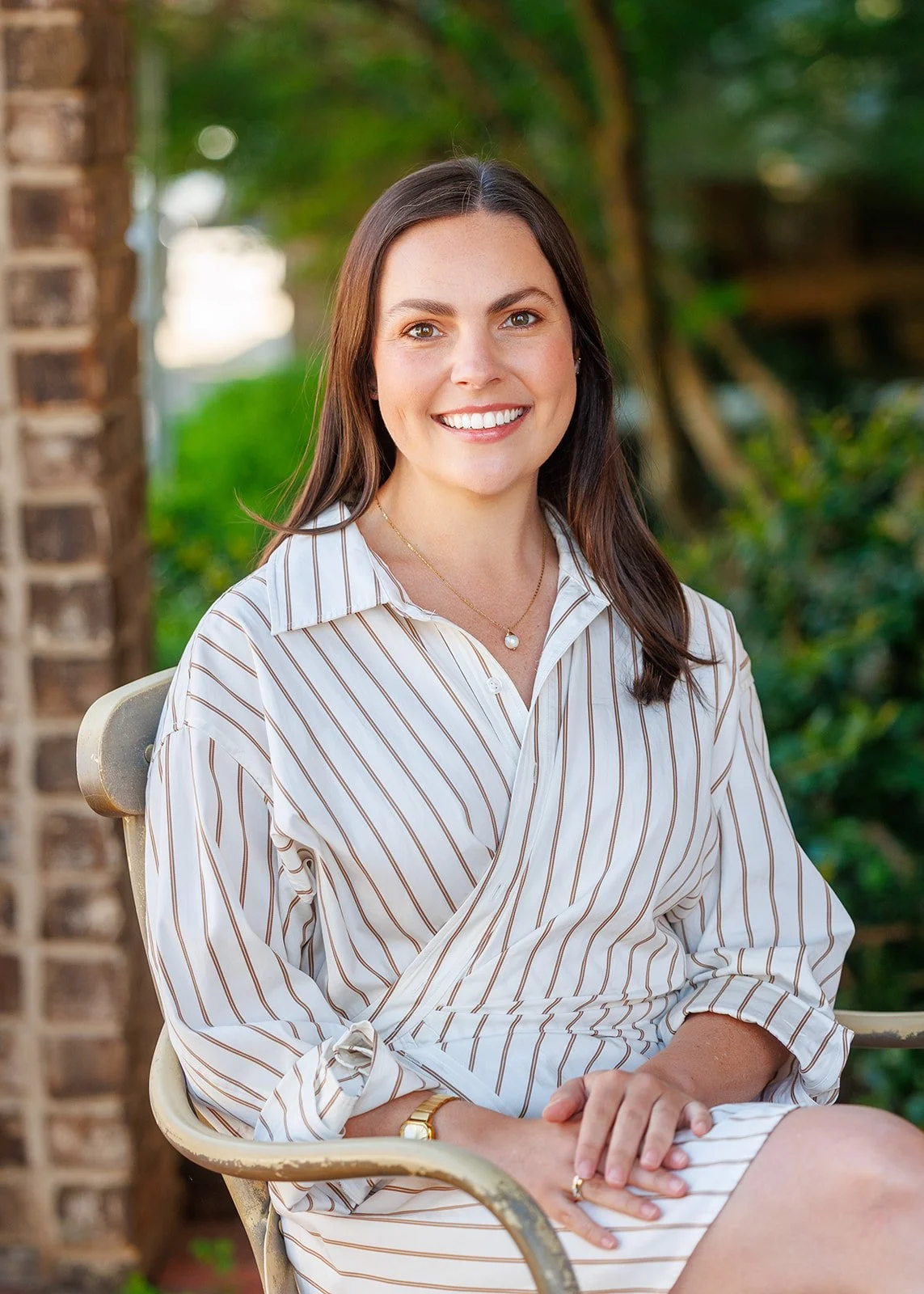 A woman with long brown hair, wearing a striped button-up dress and jewelry, sitting on a garden chair outdoors with greenery and a brick wall in the background.