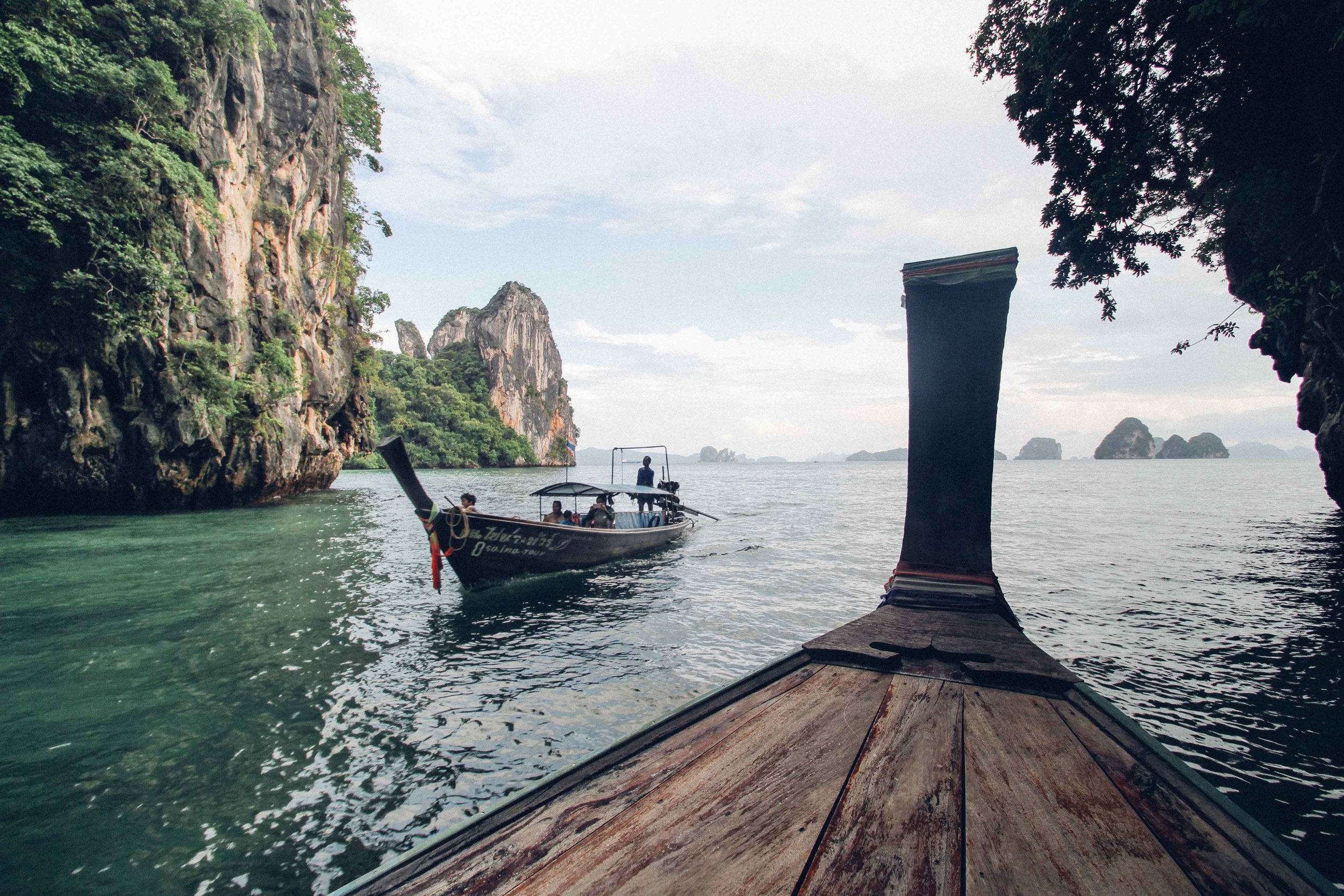 A boat with tourists floating in a bay surrounded by lush green cliffs and smaller islands, seen from a wooden dock.