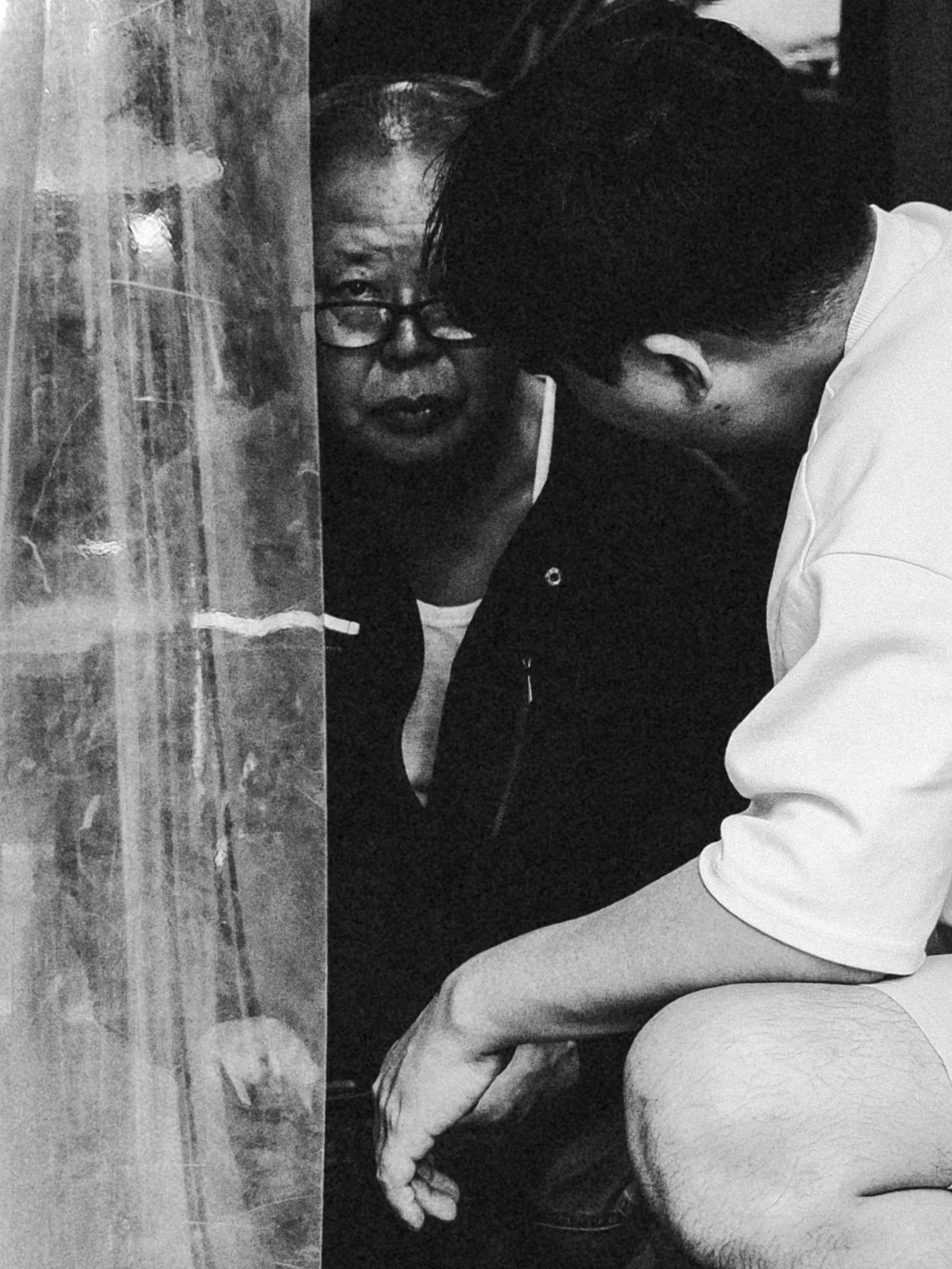 A black and white photo of a woman with glasses and her hair in a bun, sitting with her eyes looking directly at the camera. A man with short hair and wearing a white shirt is leaning towards her, and they are both near a semi-transparent curtain.