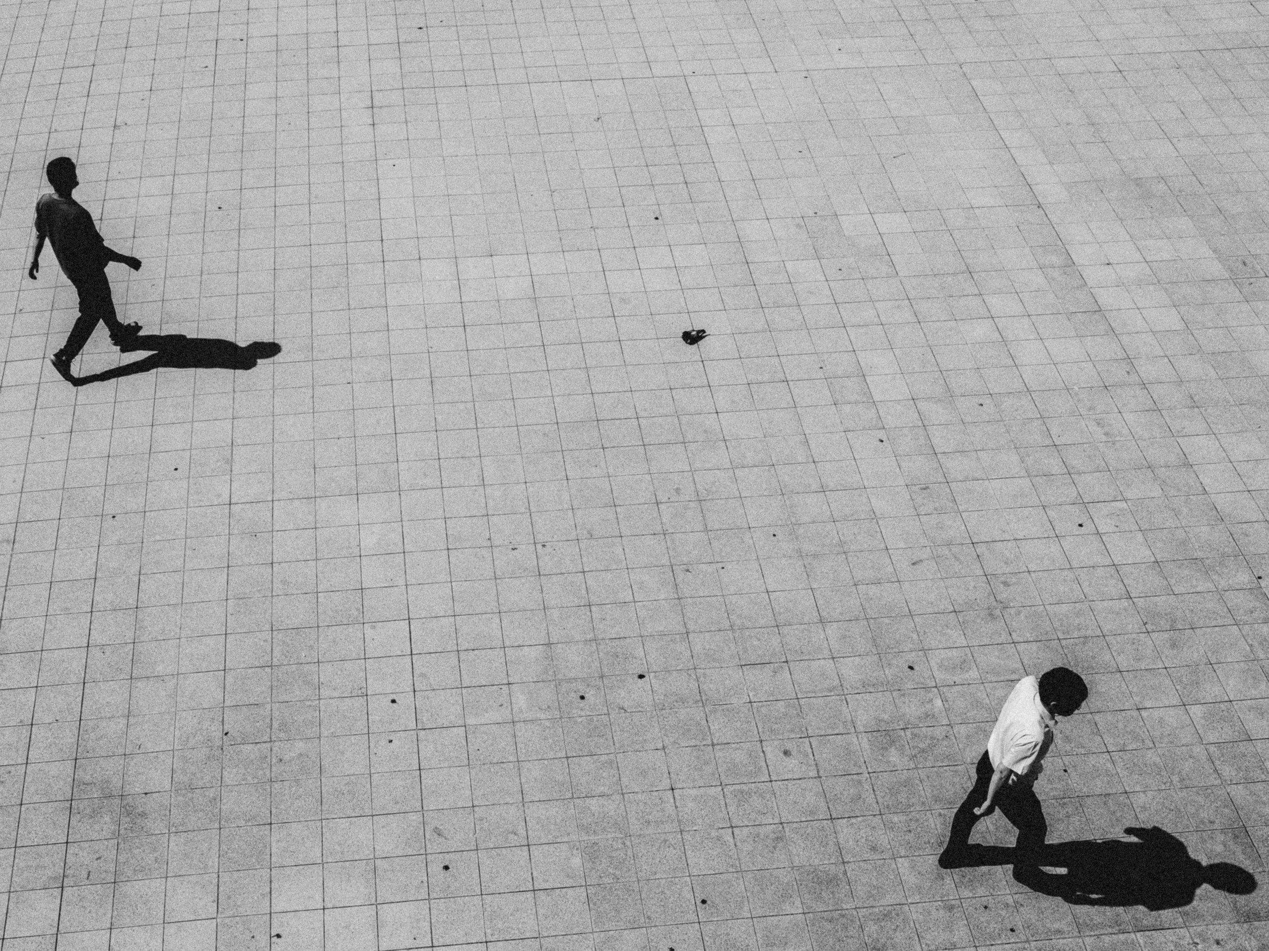 Black and white photo of two men walking on a large tiled open space. One man is near the bottom right corner, dressed in a white shirt and dark pants, walking leftward. The other man is near the upper left area, dressed in dark clothing, walking rig