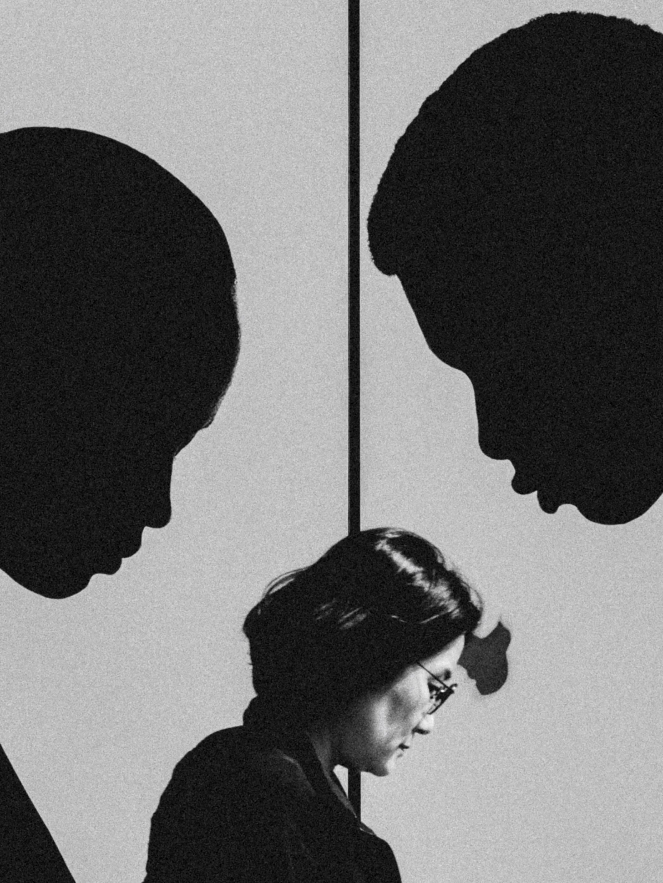 Black and white photograph of a woman wearing glasses and with her hair in a bun, looking down, with three large silhouette profiles of children’s heads in the background.