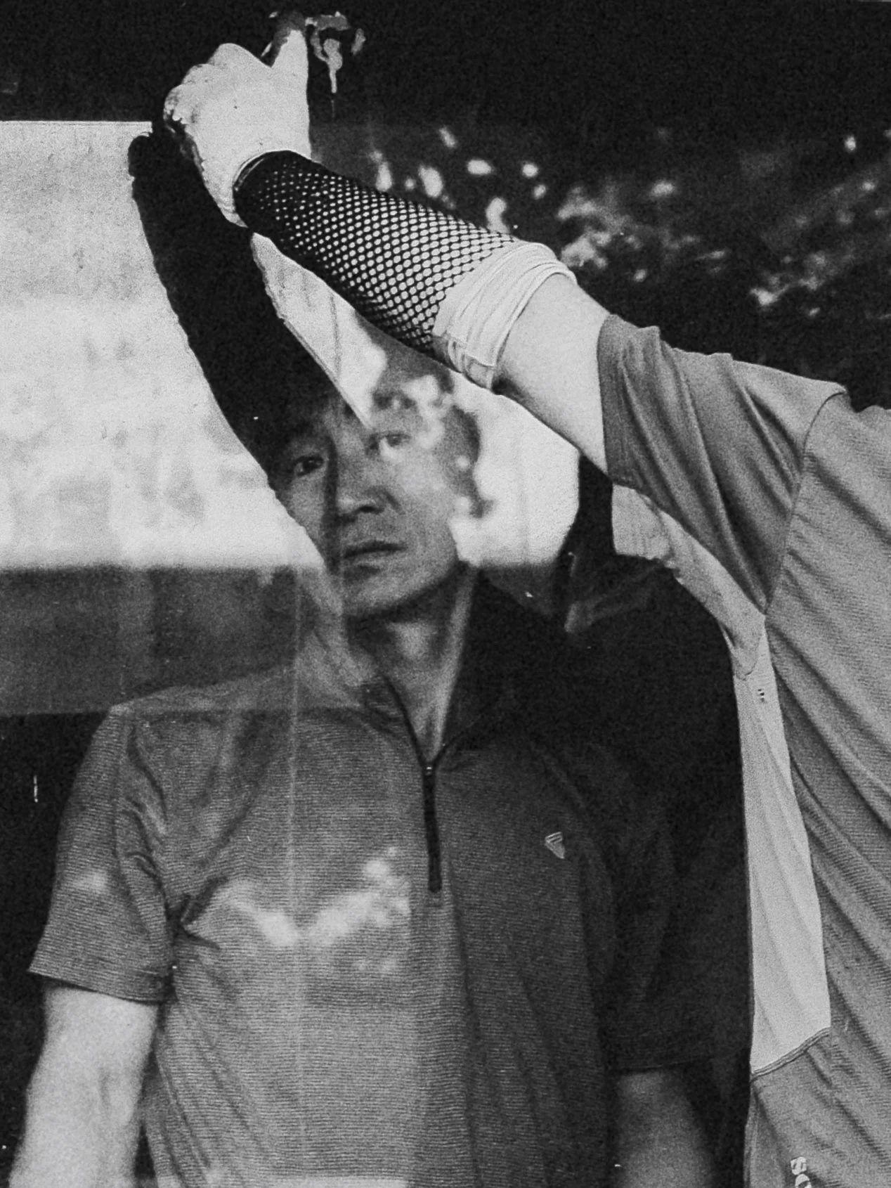 A black and white photo of two men standing outdoors at night, one man is wearing a zip-up shirt with a high collar, and the other is wearing a short-sleeved shirt with a mesh arm sleeve, with trees and lights blurred in the background.