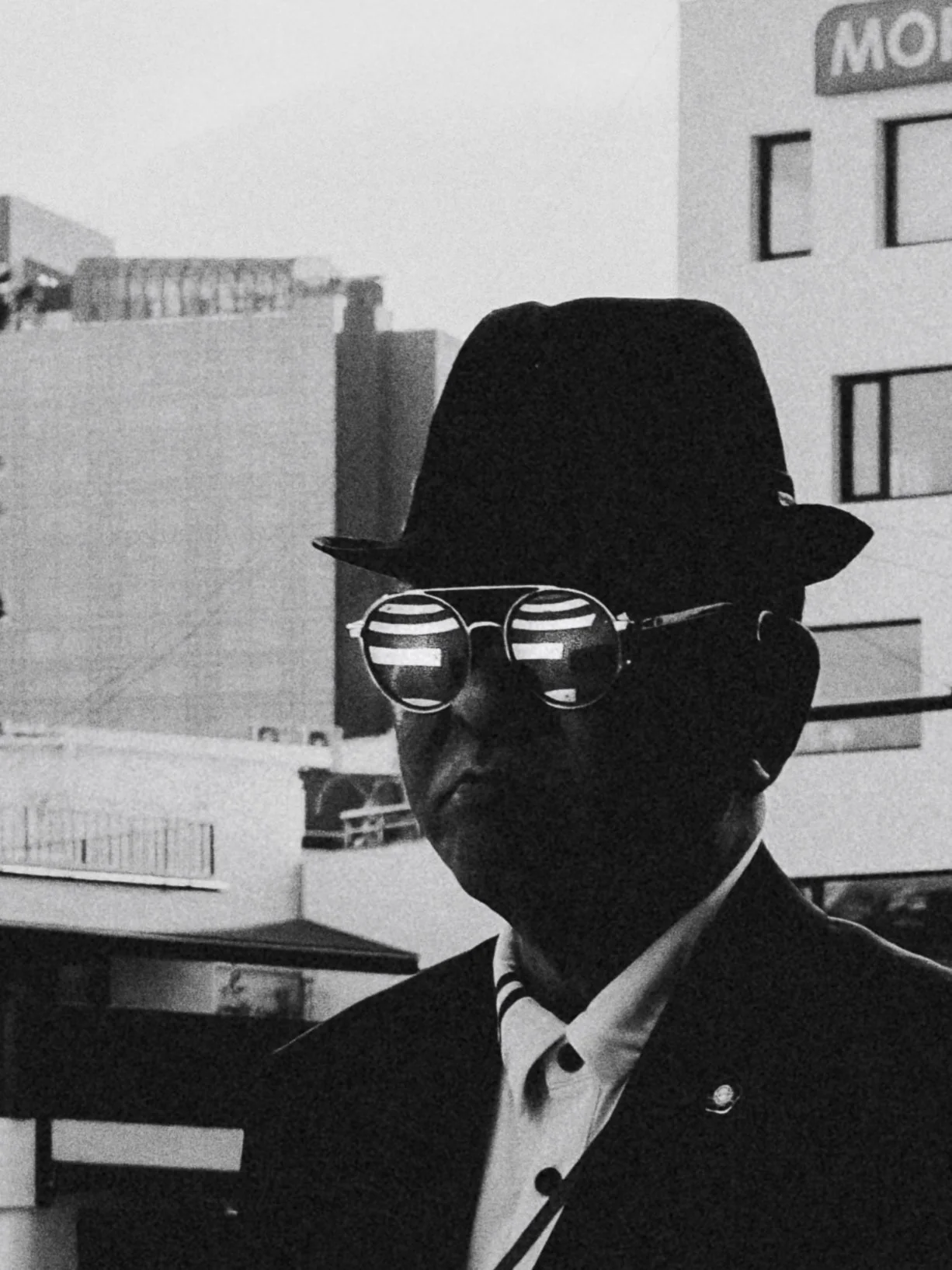 Black and white photo of a man wearing sunglasses, a hat, and a suit, standing outdoors with buildings in the background.