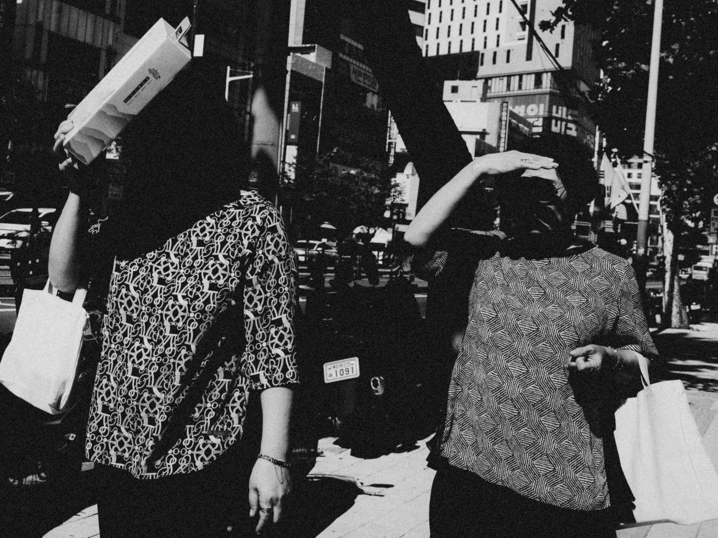 Black and white photo of two women walking outdoors in an urban setting, carrying shopping bags, with city buildings and signs in the background.