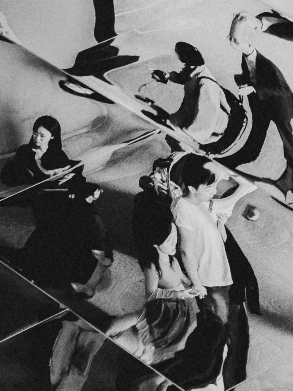 People walking on a sidewalk, some looking at their phones, in a busy urban area, reflected in glass windows.